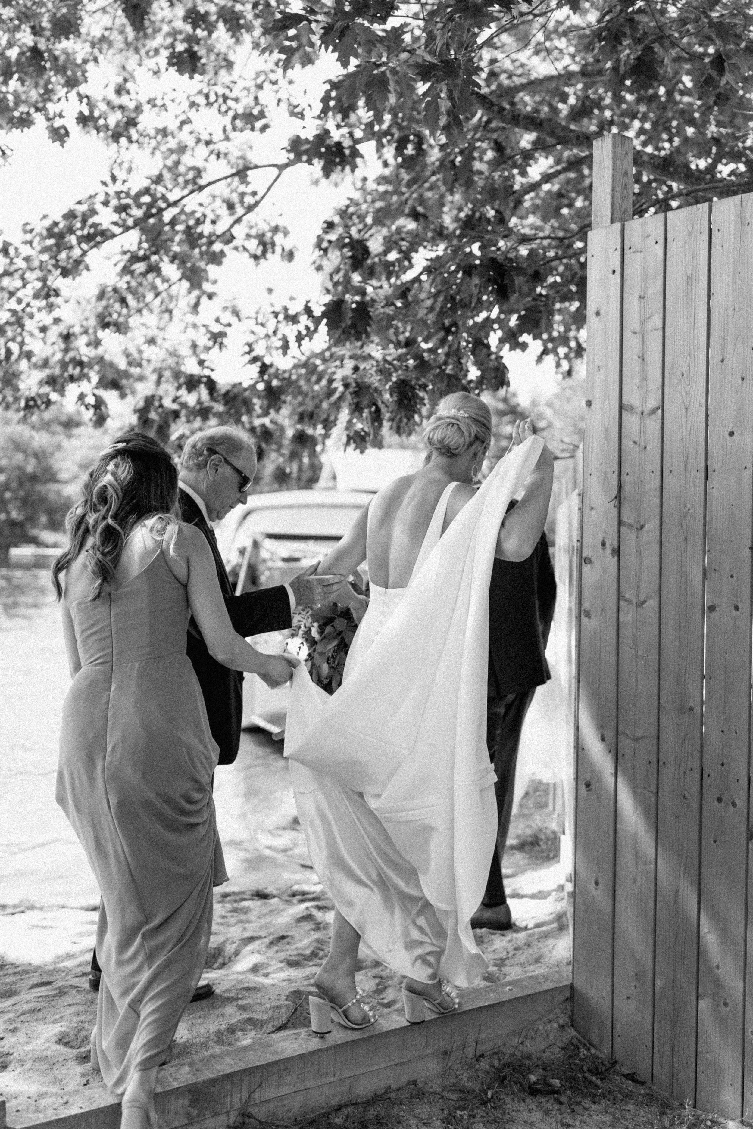A woman in a wedding dress and high heels is walking onto a beach, assisted by two people, with a wooden fence on her right and trees overhead.