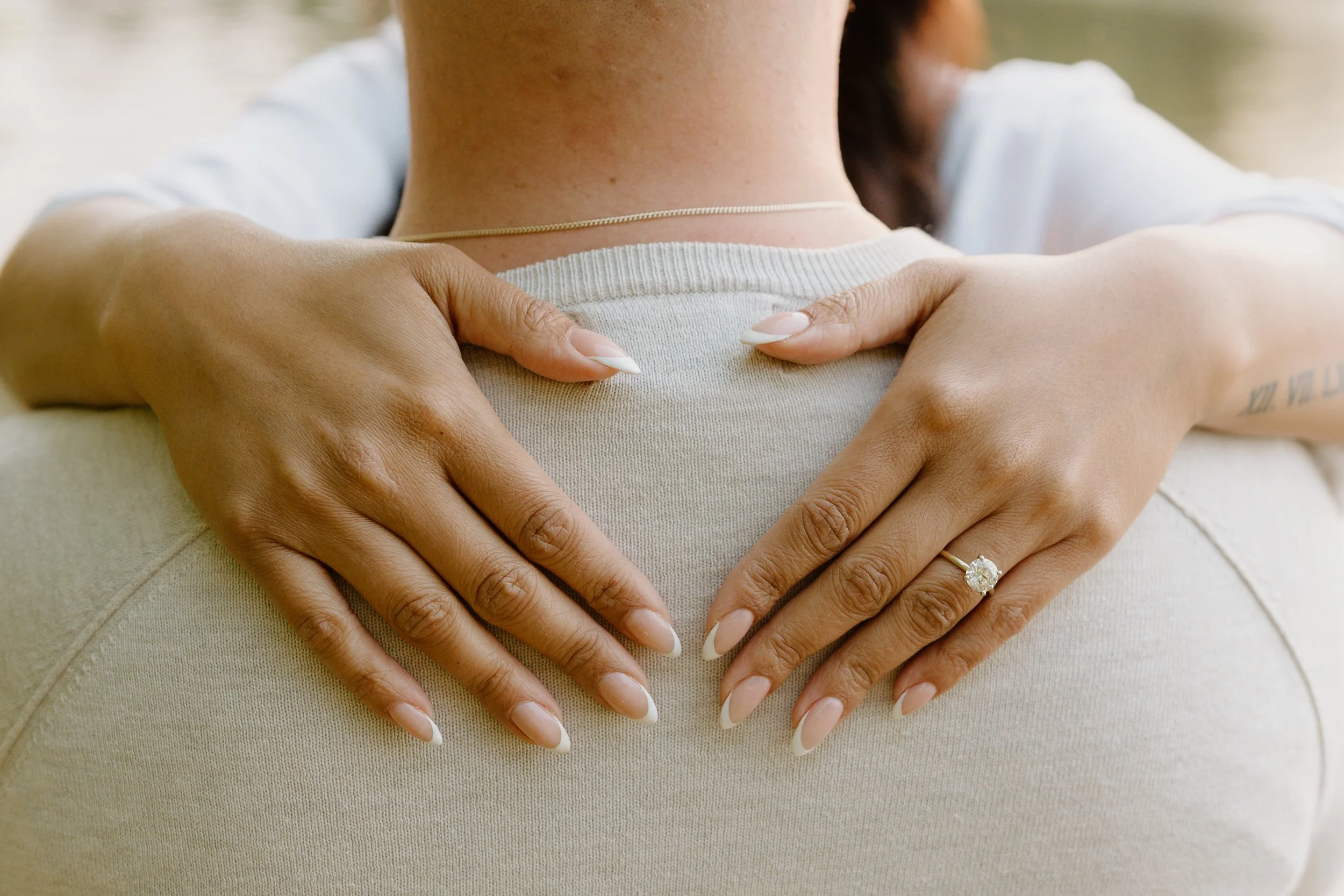 Close-up of two hands with a wedding ring embracing a person from behind, with a scenic background of a body of water.