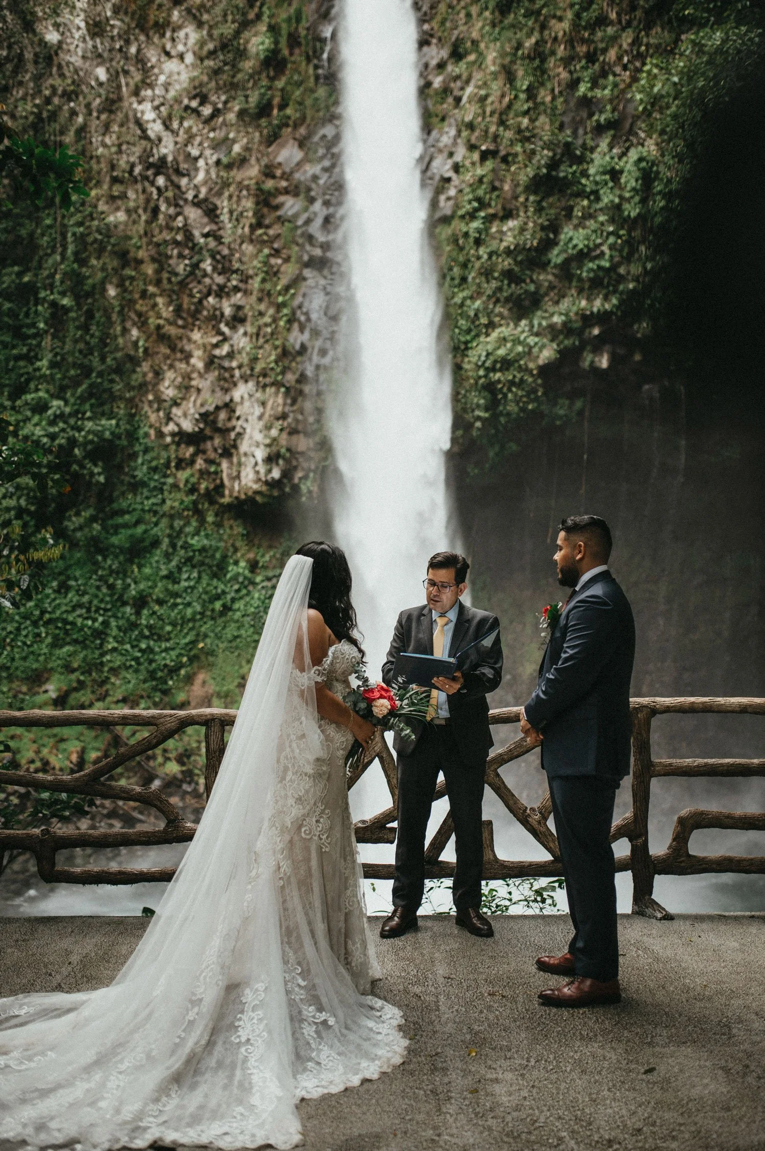 Outdoor wedding ceremony with bride in lace gown and groom in suit, officiant reading vows, set in front of a waterfall and surrounded by greenery.