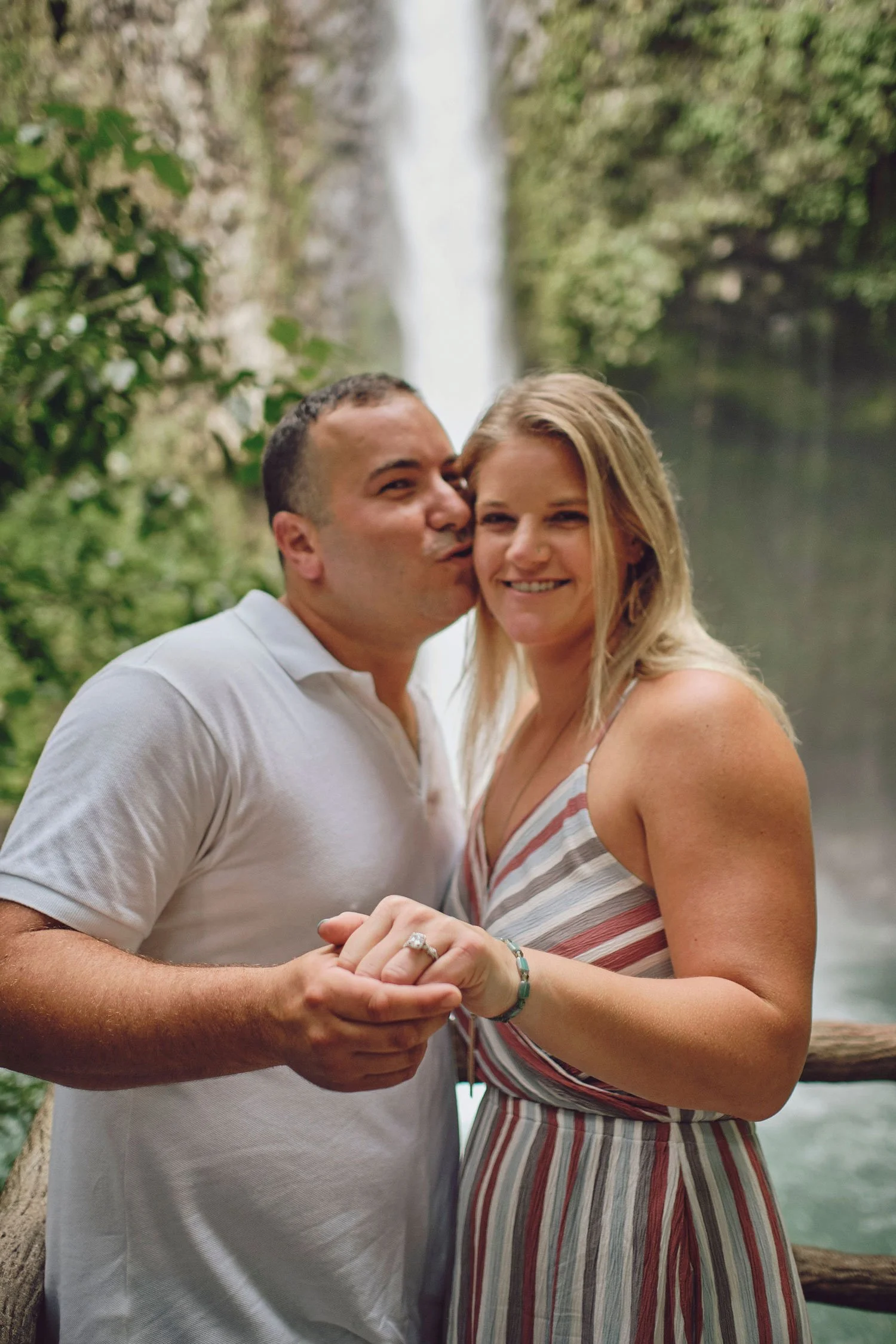 A couple posing in front of a waterfall, the man kissing the woman on the cheek while they hold hands, showing an engagement ring. The setting is outdoors with lush greenery.