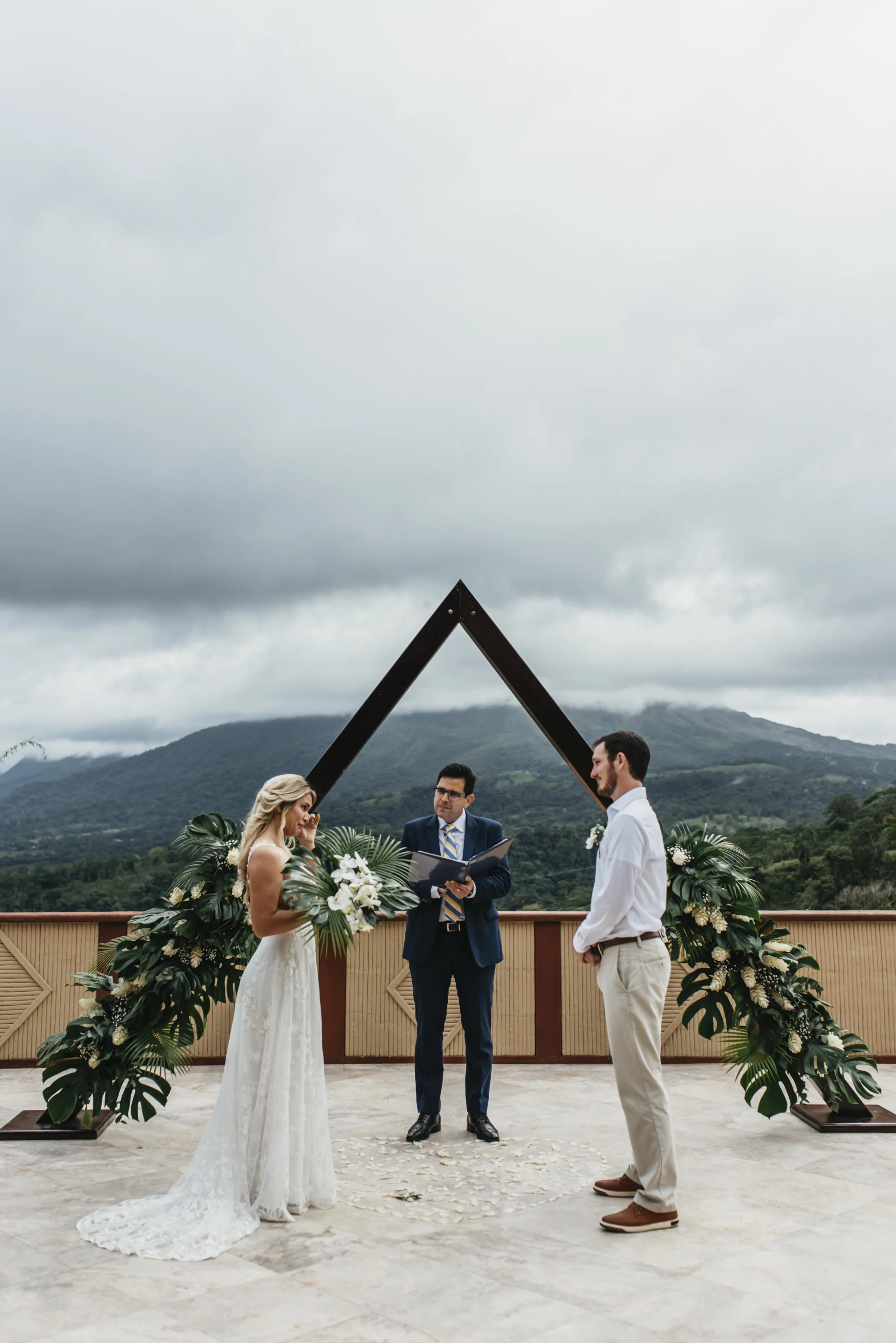 Outdoor wedding ceremony with a couple and officiant, lush greenery and mountains in the background.
