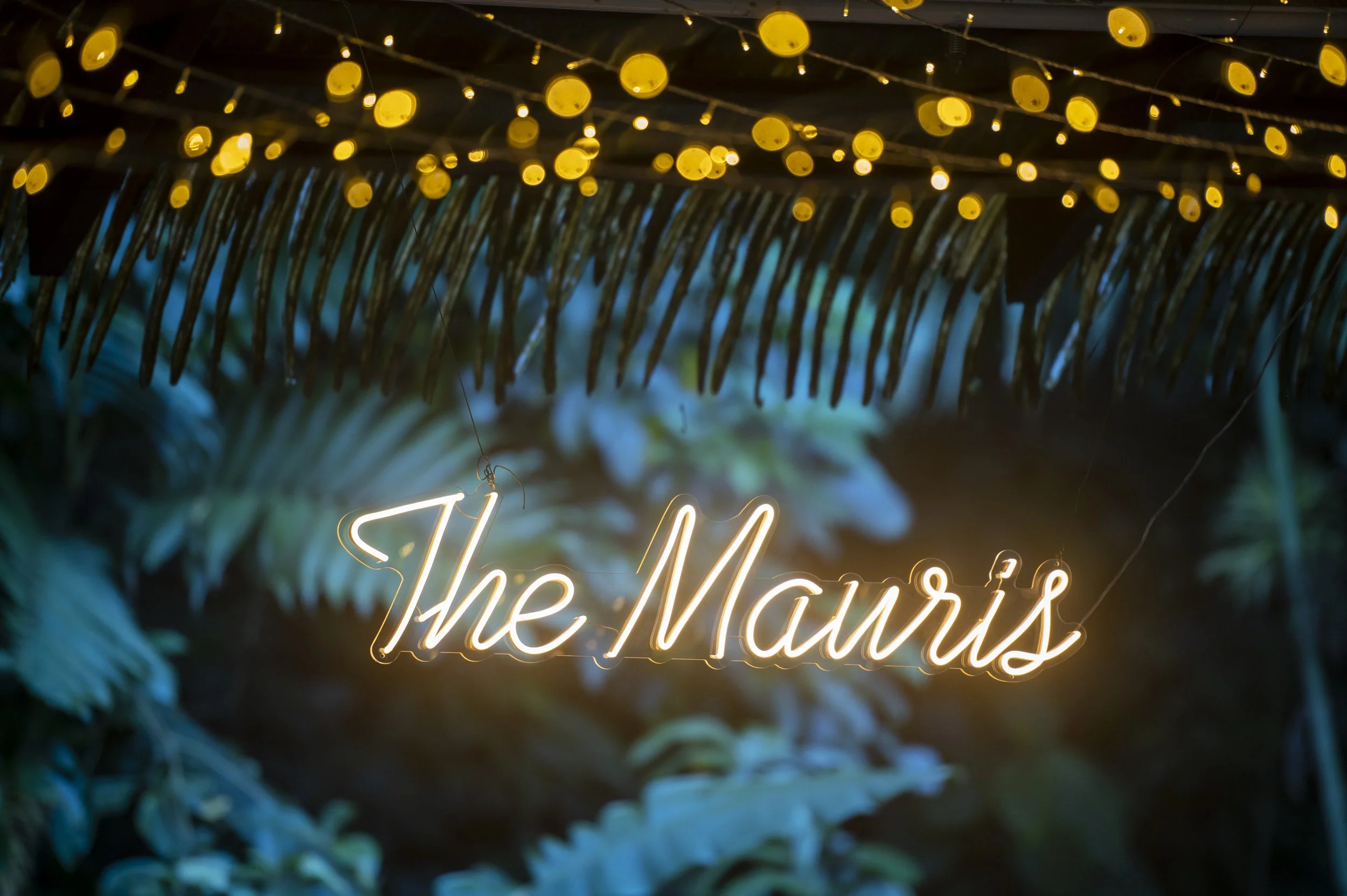 Neon sign "The Mauris" with yellow lights and tropical leaves background.