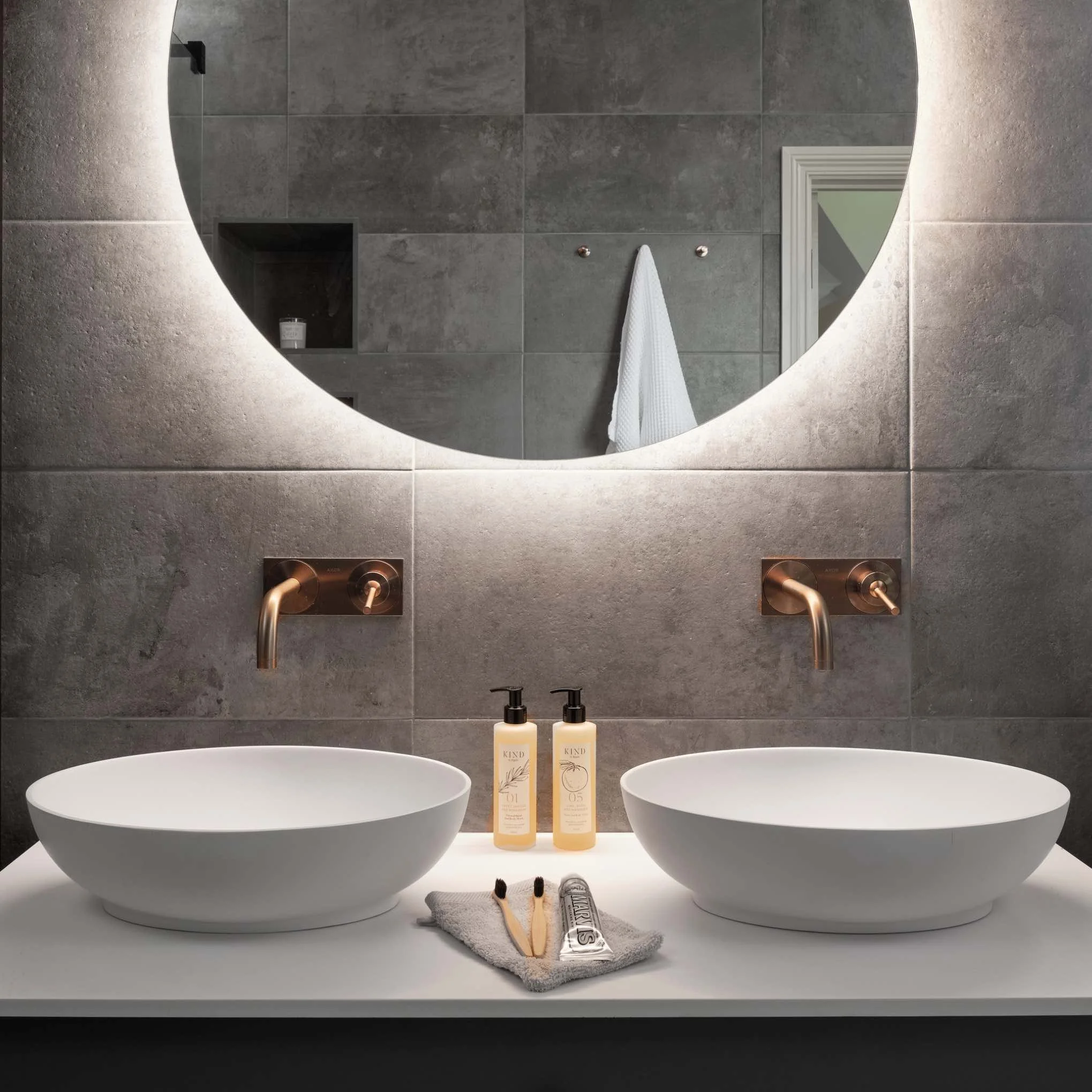 Copper basin brassware, wall mounted over two white oval basins. Large round luminated mirror above them.