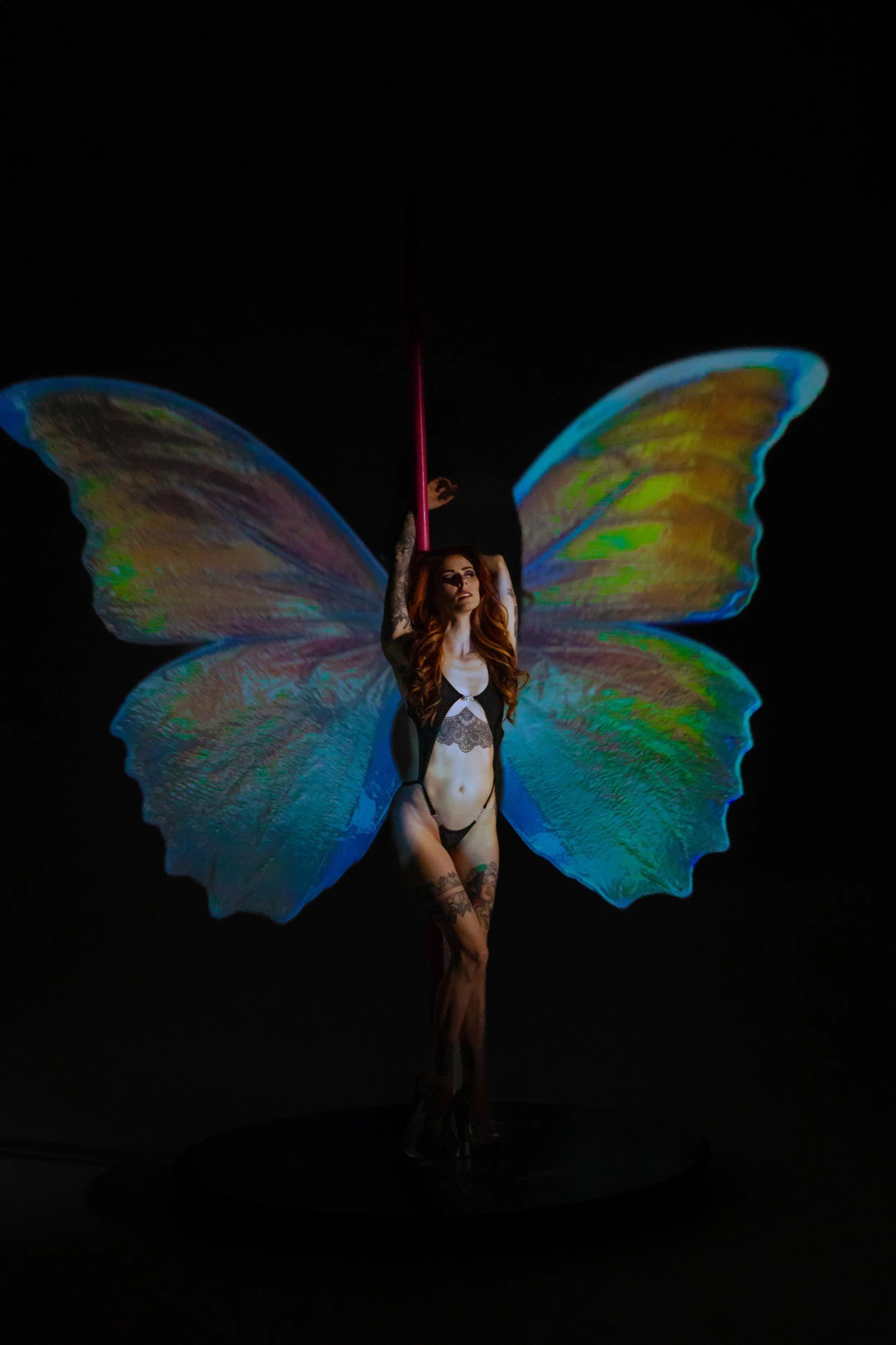 A woman with butterfly wings and tattoos is performing a pole dance against a black background.