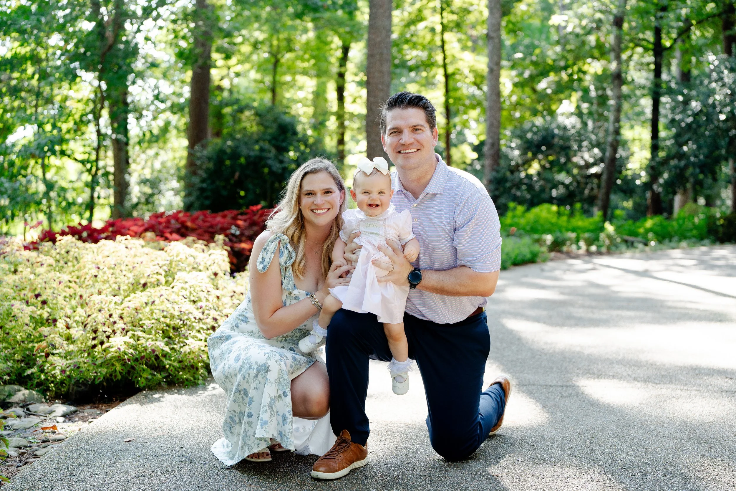 Family of three poses outdoors in a park with trees. The woman is kneeling, holding a smiling baby girl in a white dress with a bow. The man is kneeling on one knee beside them, smiling.