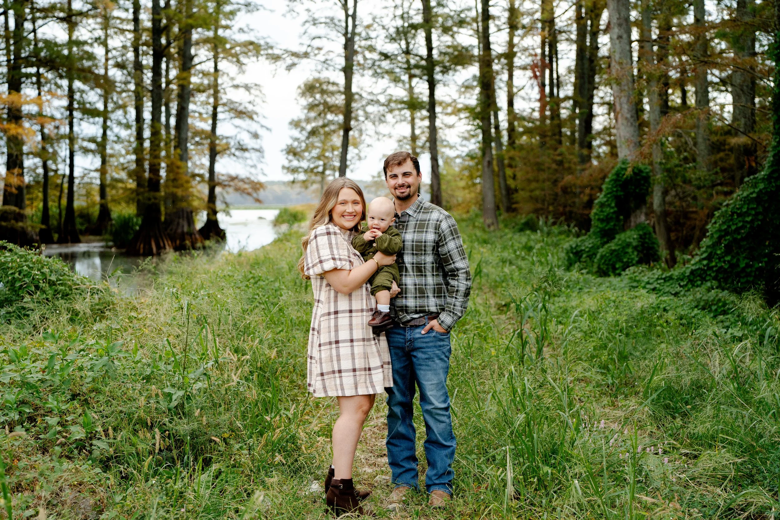 A family of three standing in a forest near a lake, smiling at the camera. The woman is holding a baby boy, and the man stands beside them with one hand in his pocket. They are dressed in plaid shirts, surrounded by green grass and trees.