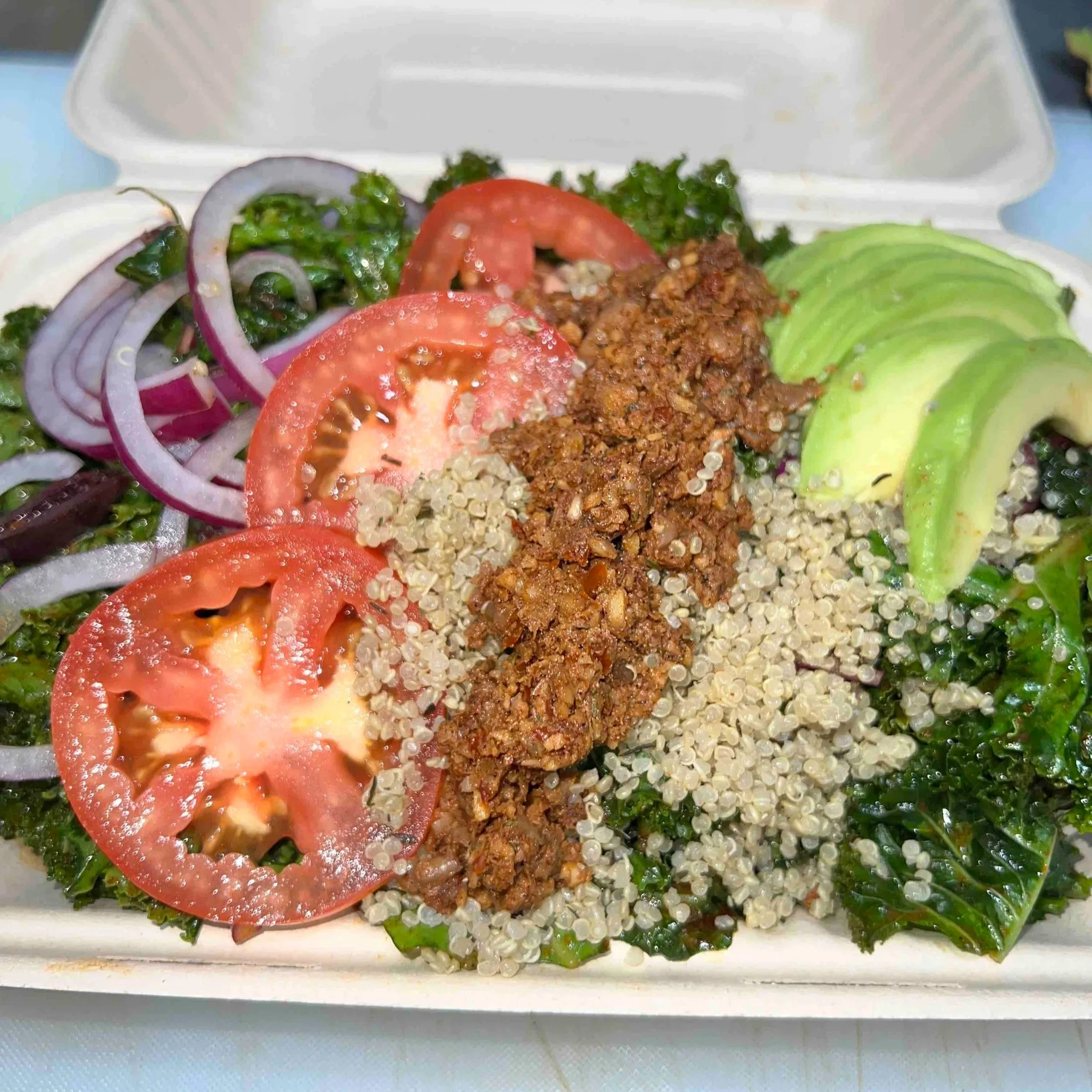 Salad with tomatoes, onions, avocado, quinoa, kale, and seasoned ground beef in a takeout container