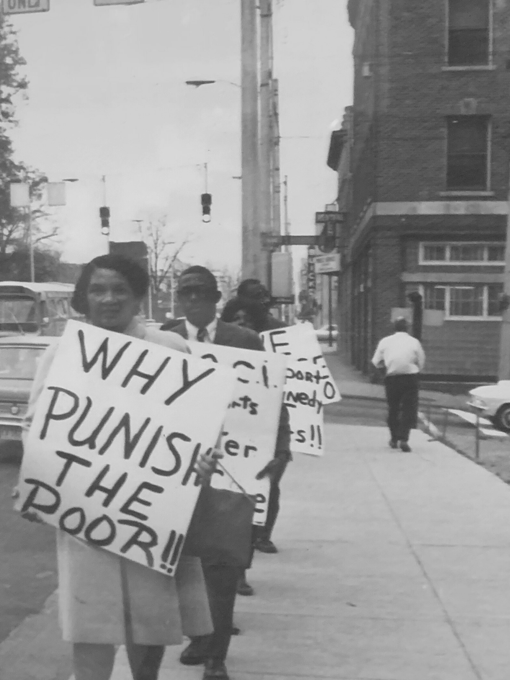 black and white photo of people protesting, woman in front holds a sign saying "why punish the poor?"