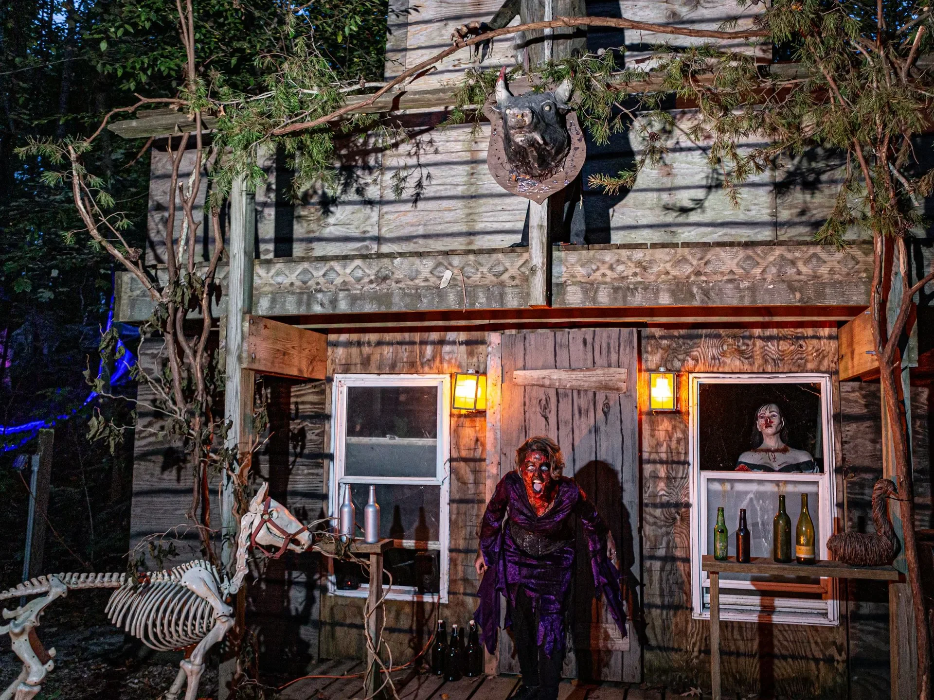 A haunted house scene with two women dressed as zombies, one with face paint and the other with a skeleton mask, standing outside a wooden house decorated with eerie figures, skeletons, and dim lights.