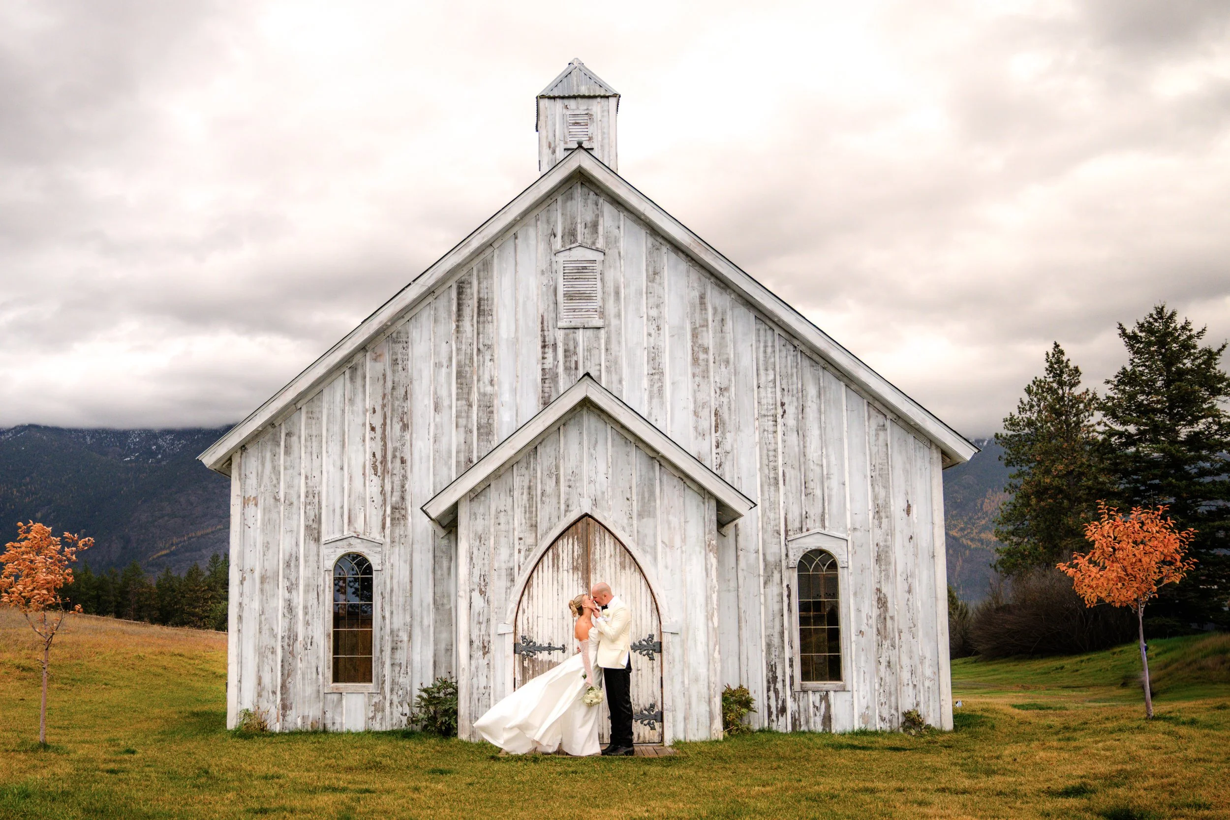 A bride and groom kissing in front of a weathered white wooden church with mountains and cloudy sky in the background.