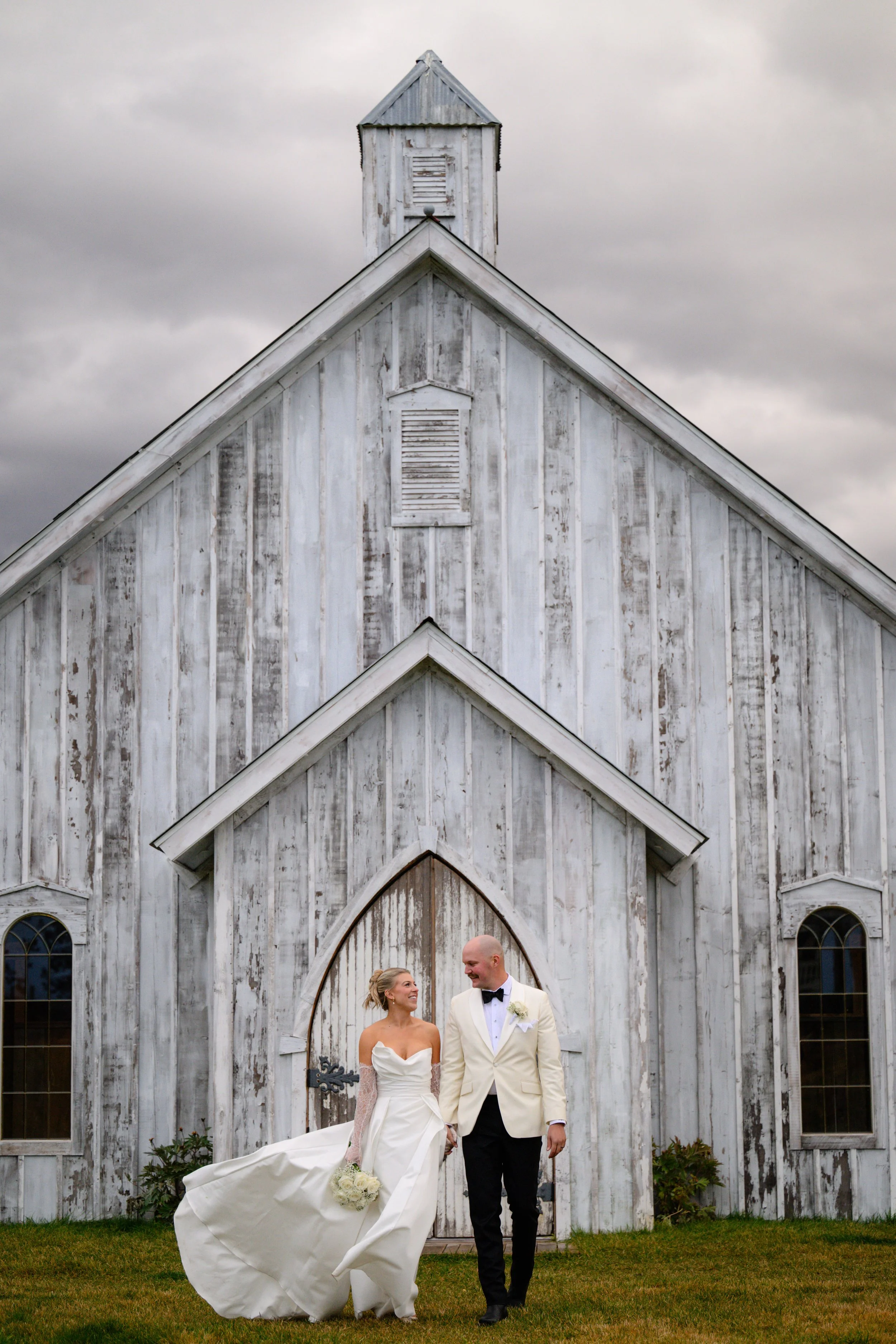 A newlywed couple holding hands and smiling while walking in front of a rustic white church with a peaked roof and small steeple, under a cloudy sky.