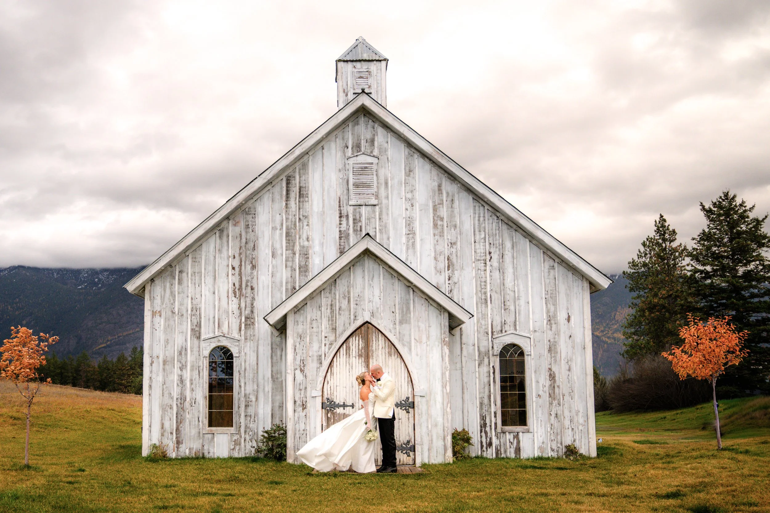 A Montana white wedding chapel venue near Glacier National Park. A bride and groom kissing in front of a rustic, white wooden church with gothic windows, surrounded by a grassy field with autumn trees, mountains in the background, and a cloudy sky.