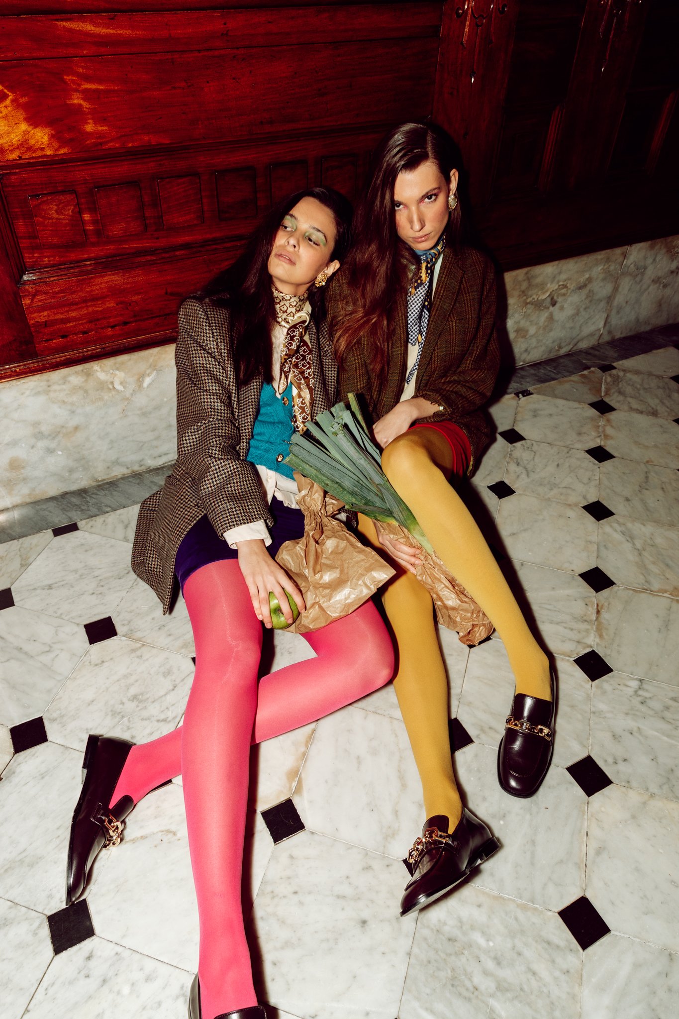 Two stylishly dressed women sitting on a marble floor, wearing colorful tights and loafers, holding a paper bag with leeks and an apple.