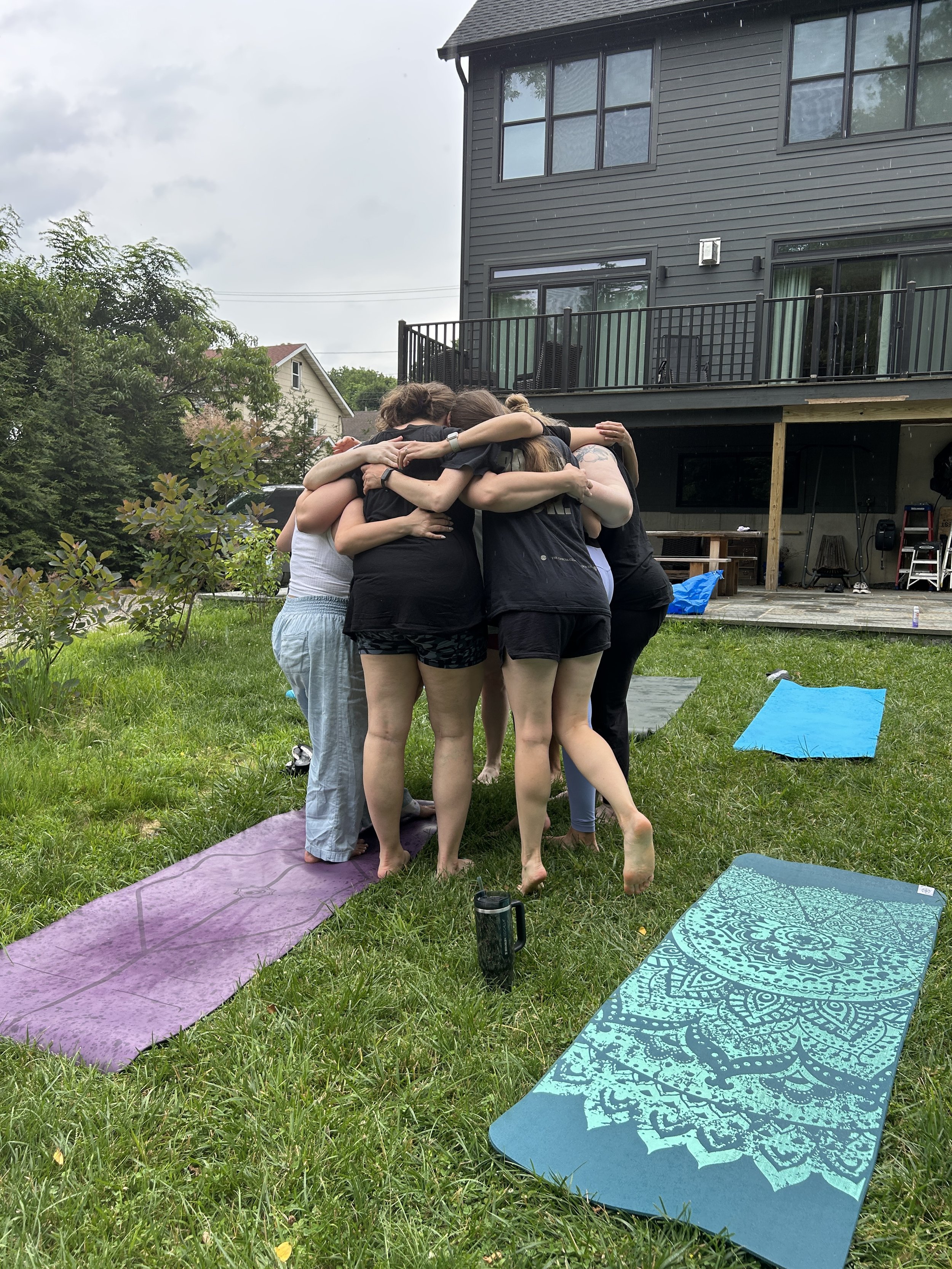 Group of people hugging outdoors on a grassy lawn with yoga mats and a house in the background.