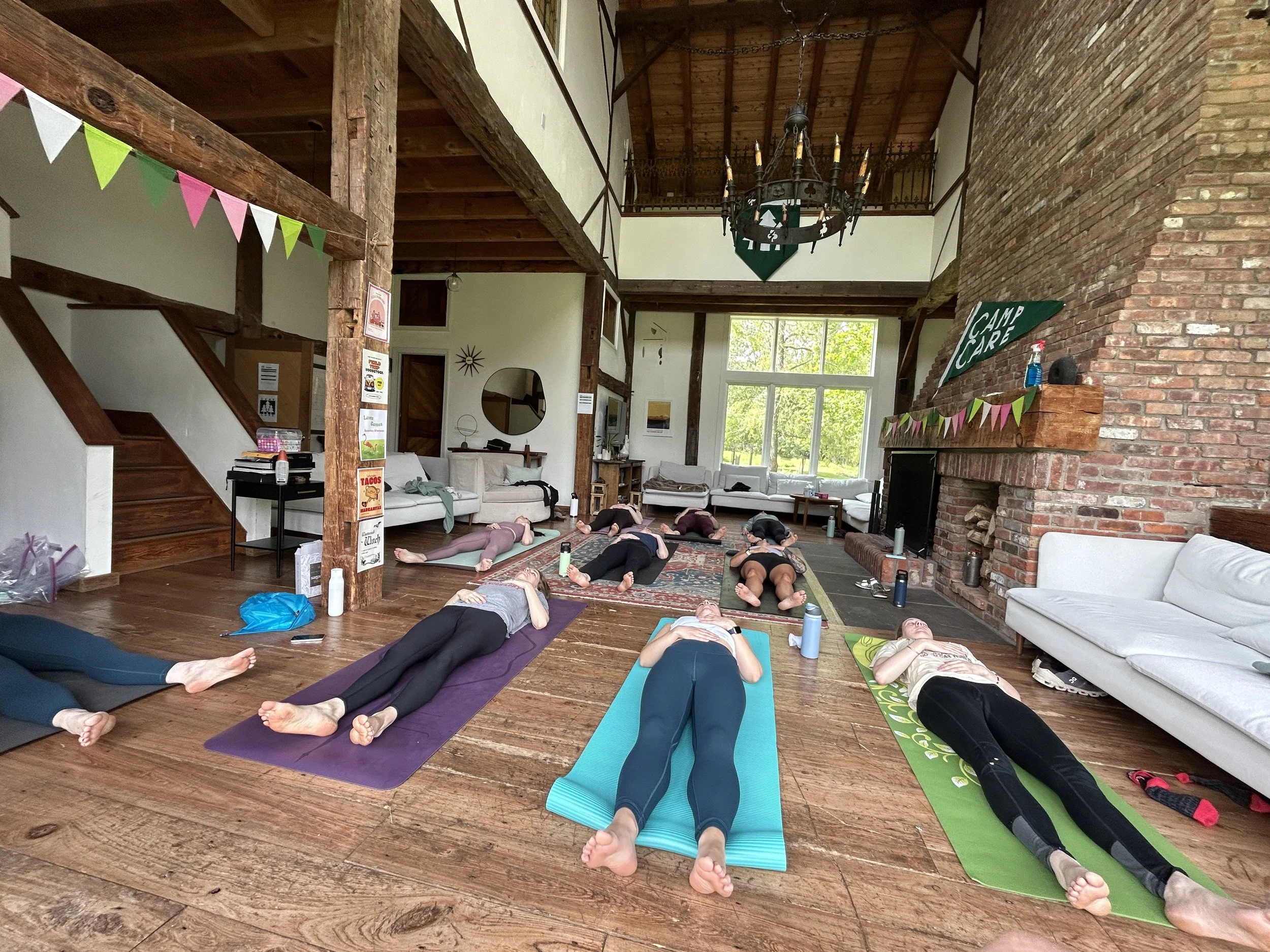 A group of people participating in a yoga class inside a spacious living room with wooden beams, brick fireplace, and large windows, lying on yoga mats in a supported pose.