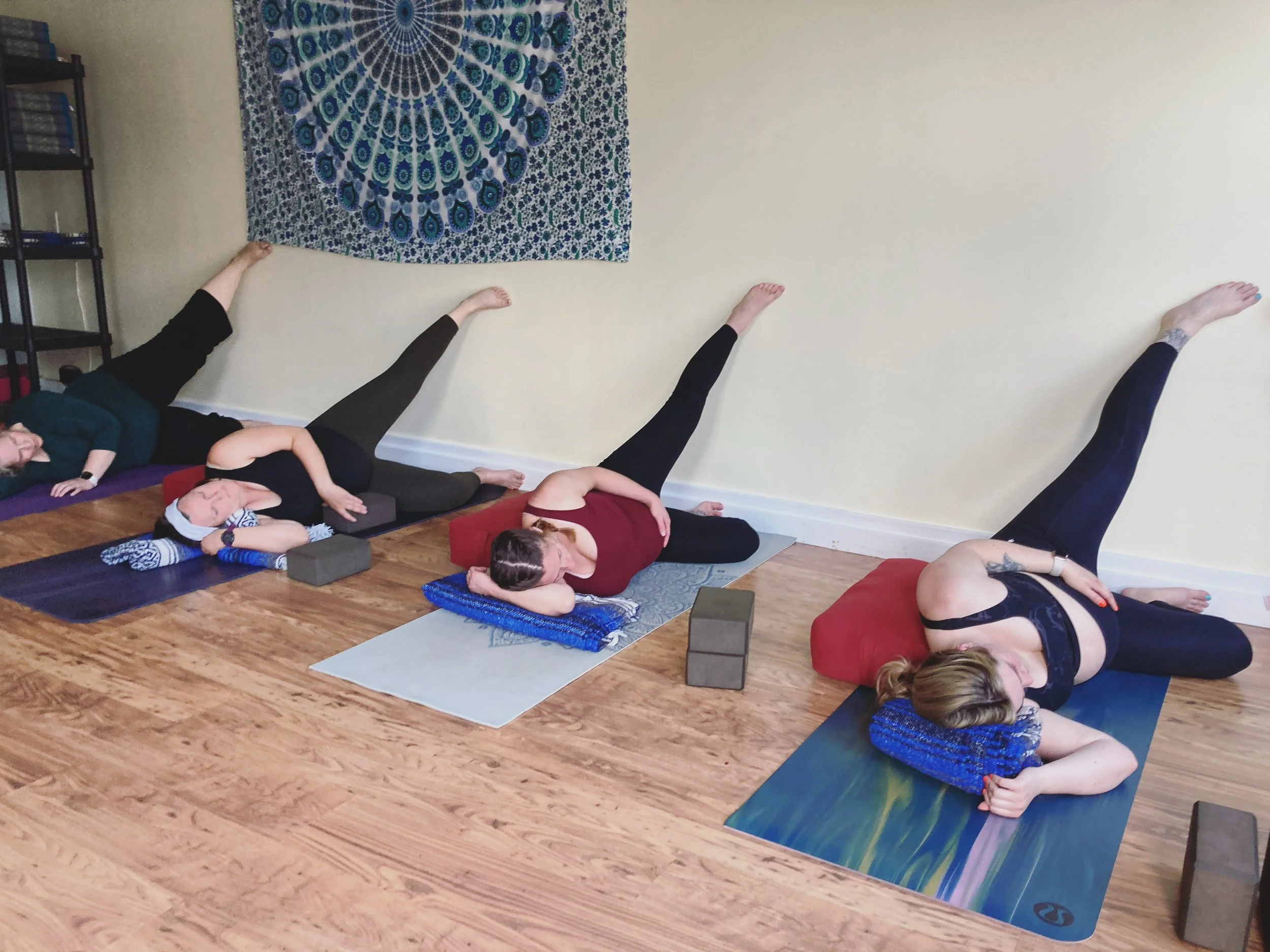 A group of pregnant people practicing yoga indoors, lying on yoga mats with their legs raised against the wall.