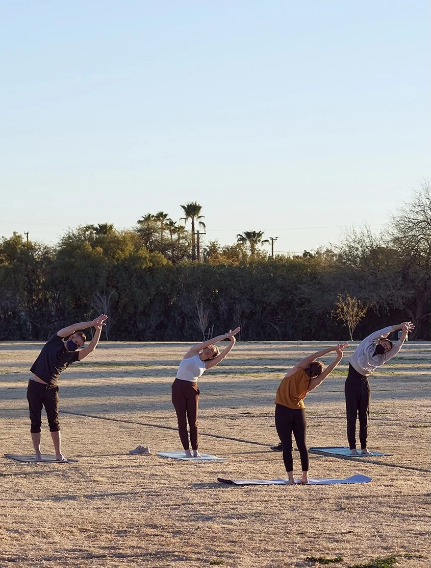 Four people practicing yoga outdoors on mats in a sandy field during sunset, with trees and power lines in the background.