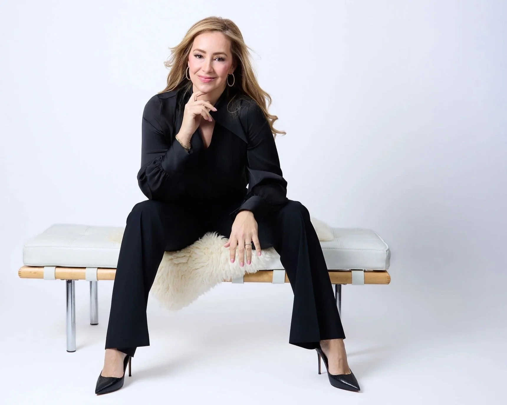 A woman in a black outfit sitting on a bench with a white furry throw, smiling and posing with her hand near her chin against a plain white background.