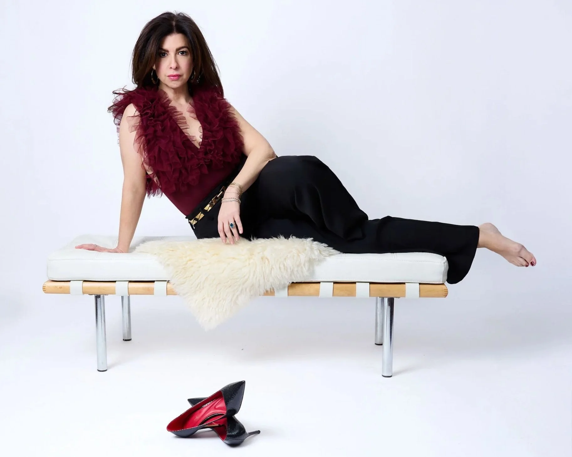 A woman lying on a white bench with a cream-colored fur rug, wearing a burgundy ruffled top and black pants, with a pair of black and red high heels on the floor nearby.