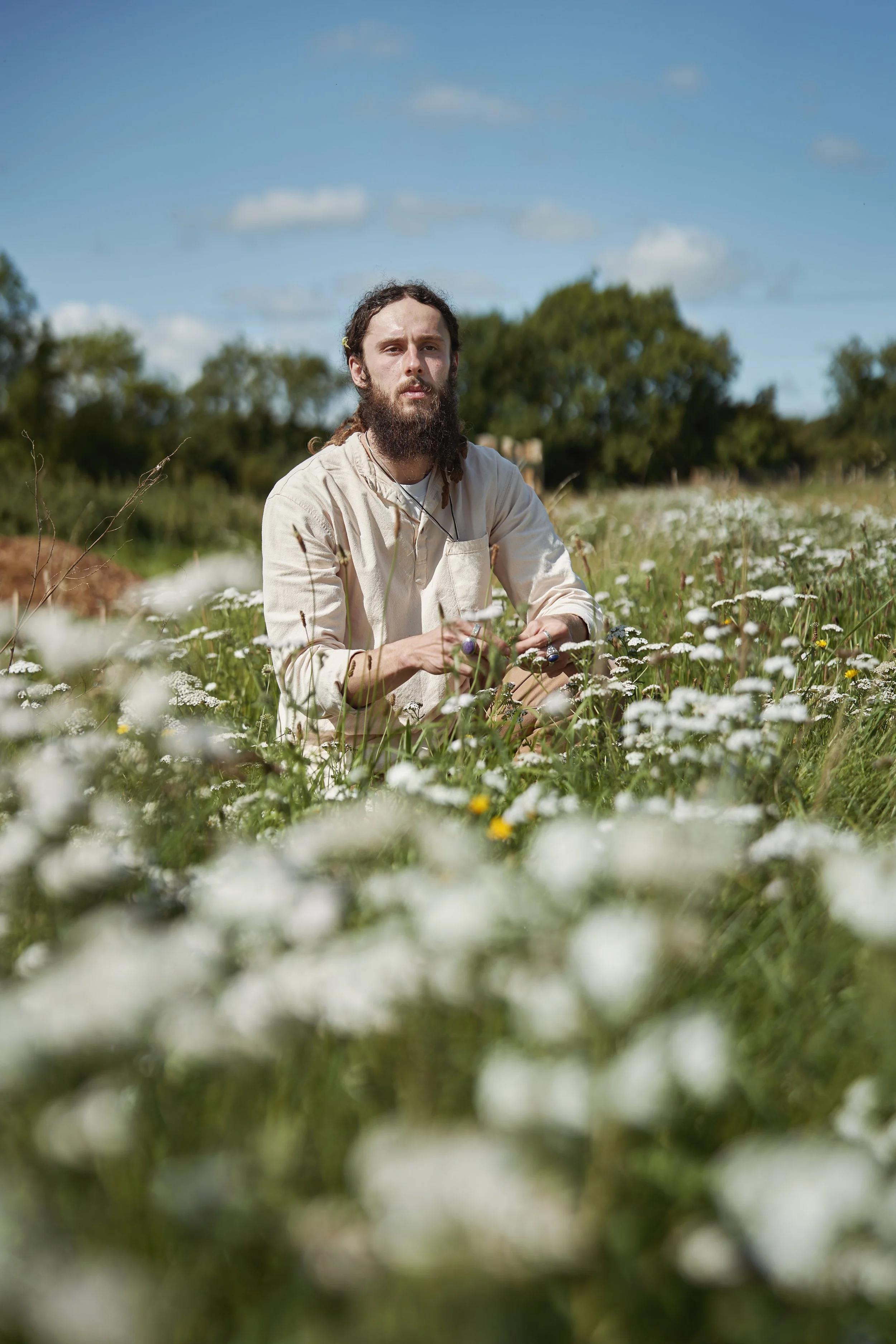A man with a beard and long hair in a field of white Yarrow flowers on a sunny day.