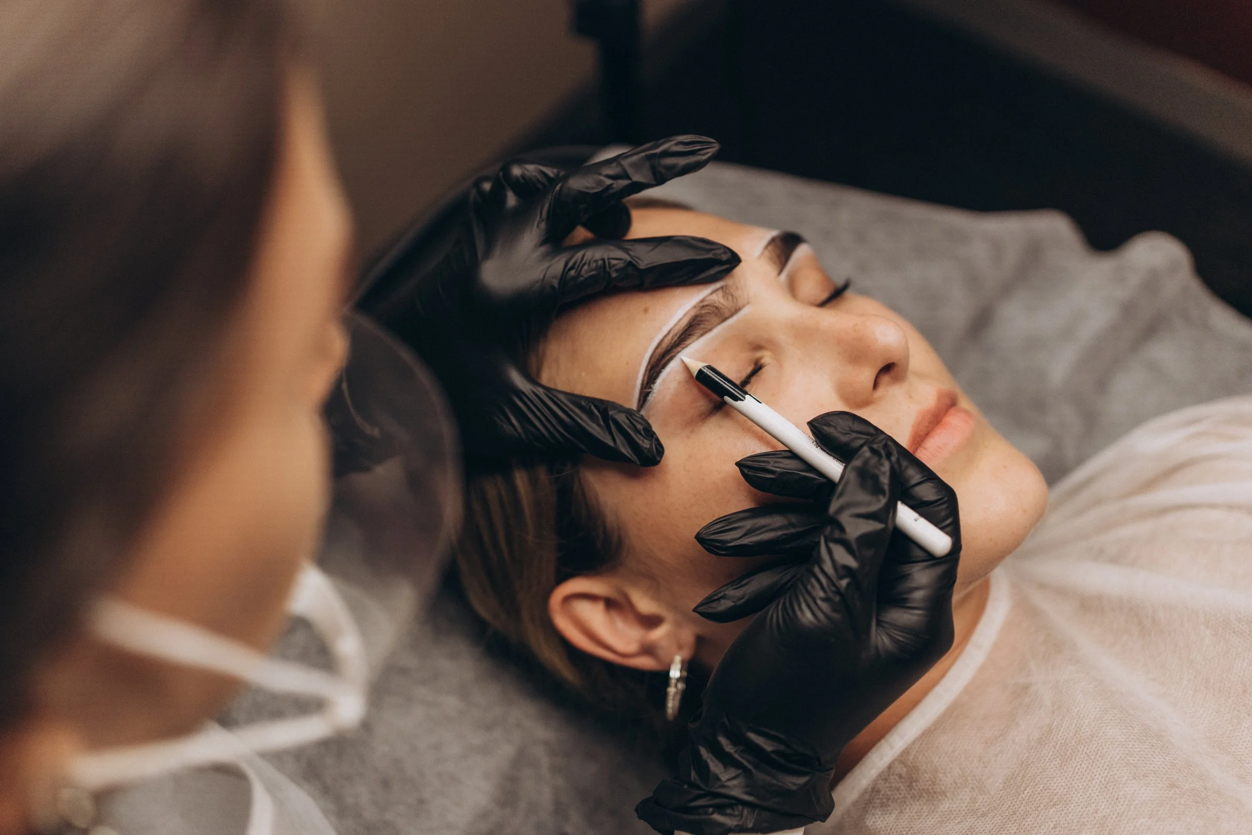 A woman is lying down with her eyes closed as a beauty technician sketches on her eyebrows with a black marker, wearing black gloves.