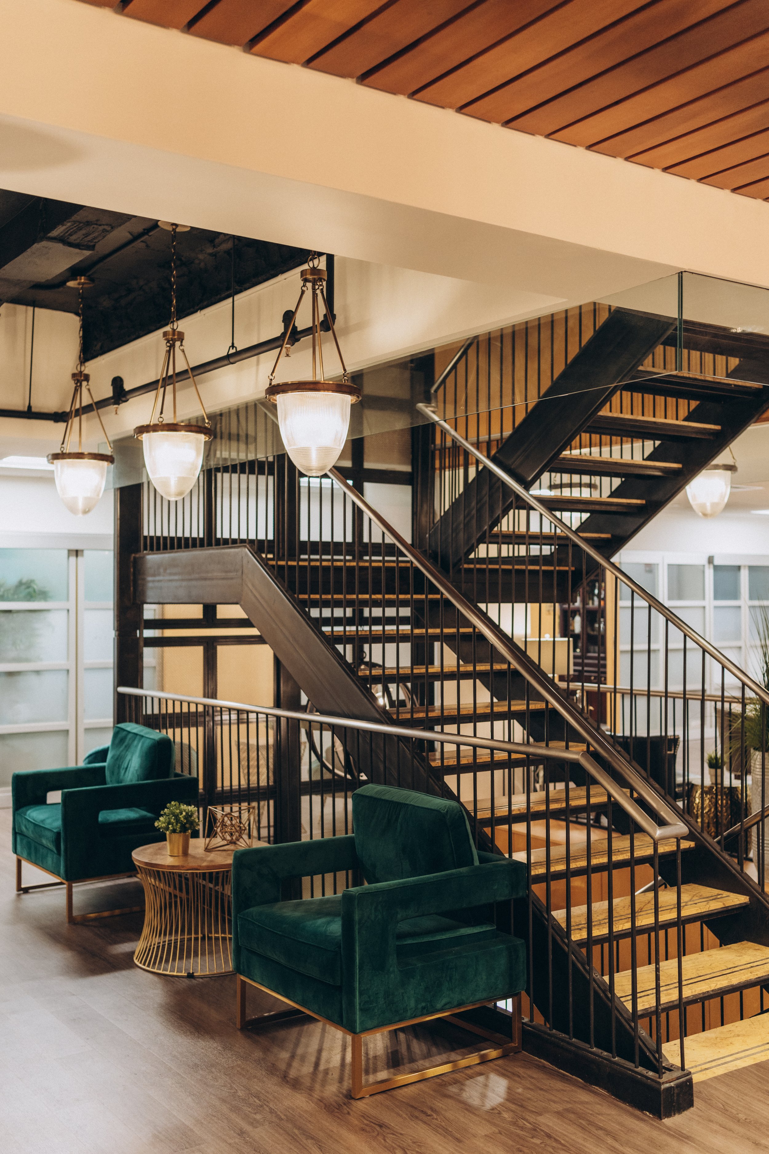 Interior view of a modern lobby with green armchairs, a small wooden table with a plant, a staircase with wooden steps and black railing, and hanging light fixtures.
