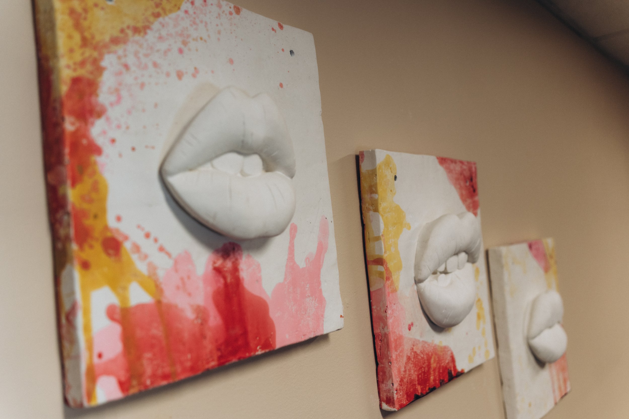 Three abstract paintings of lips on canvas, featuring white lips with paint splashes of yellow, red, and pink, displayed on a beige wall.