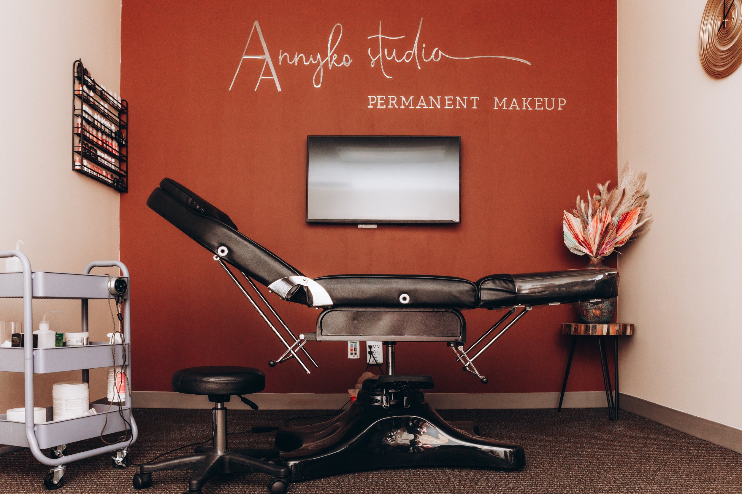 Beauty treatment room with a reclining chair, a flat-screen TV on a red accent wall with Microsoft Word writing 'Annylka studio' and 'PERMANENT MAKEUP,' a cart with beauty supplies, and a decorative plant on a side table.