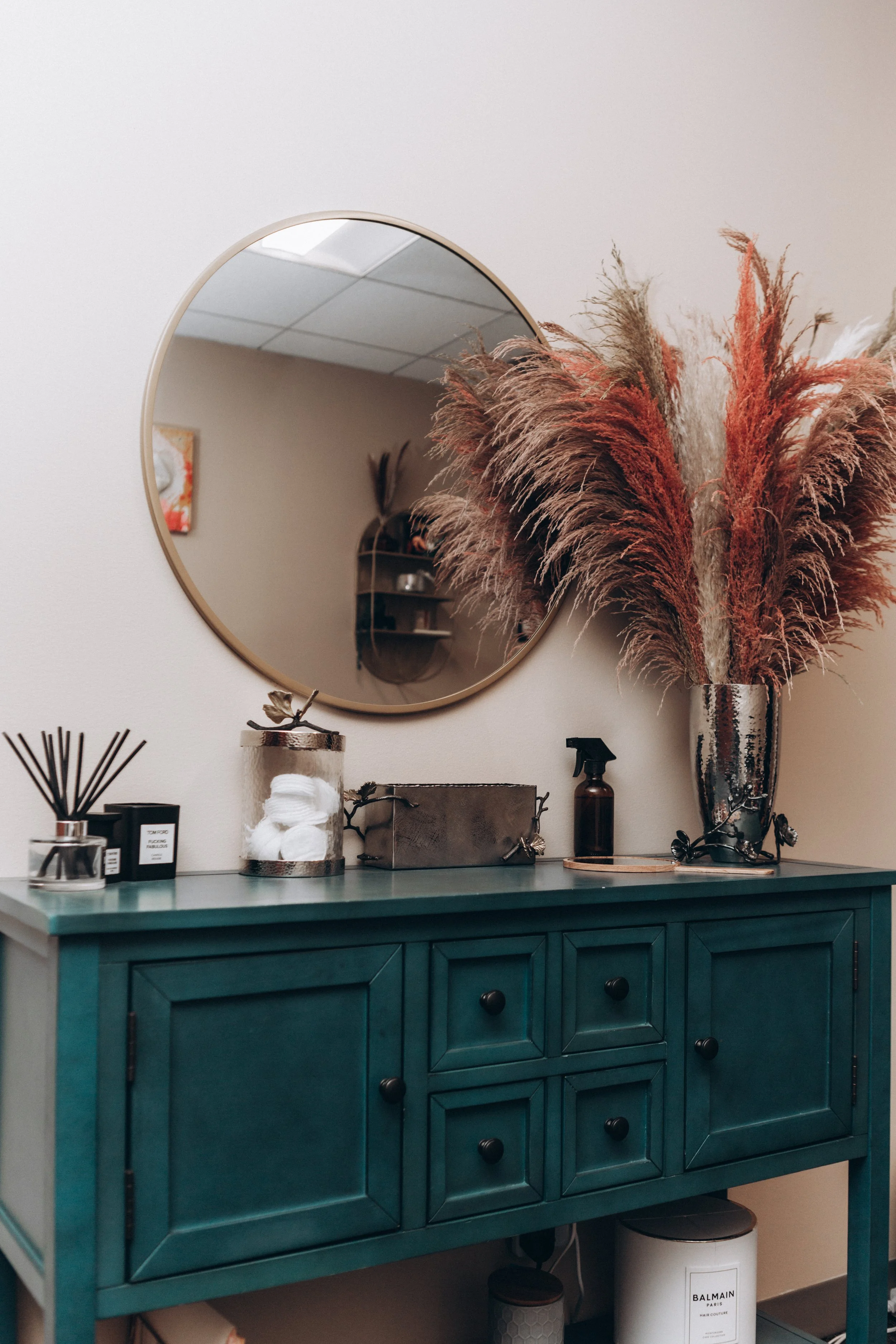 Decorative hallway or bathroom vanity with a teal cabinet, a round mirror, a tall vase with dried pampas grass, bottles, and a box of cotton balls.
