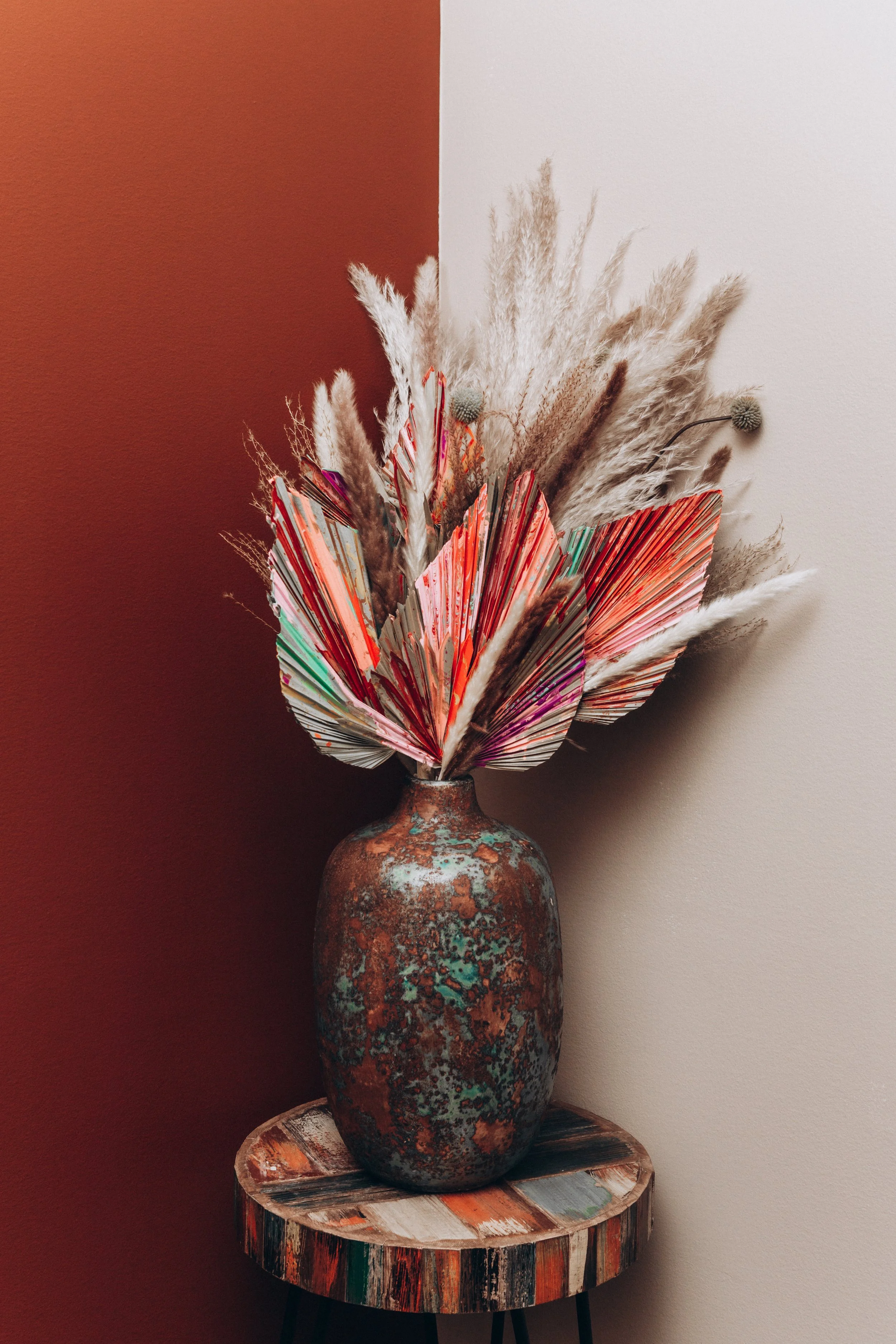 A rusted, multicolored ceramic vase with a textured surface holds an arrangement of dried pampas grass, decorative dried flowers, and colorful fan-shaped paper or fabric accents, placed on a wooden side table against a two-tone wall.