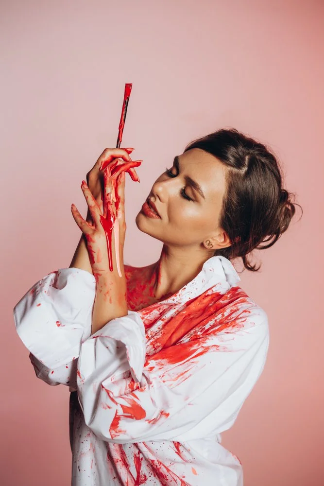 A woman with makeup and styled hair, wearing a white shirt covered in red paint, holding a paintbrush near her face against a pink background.