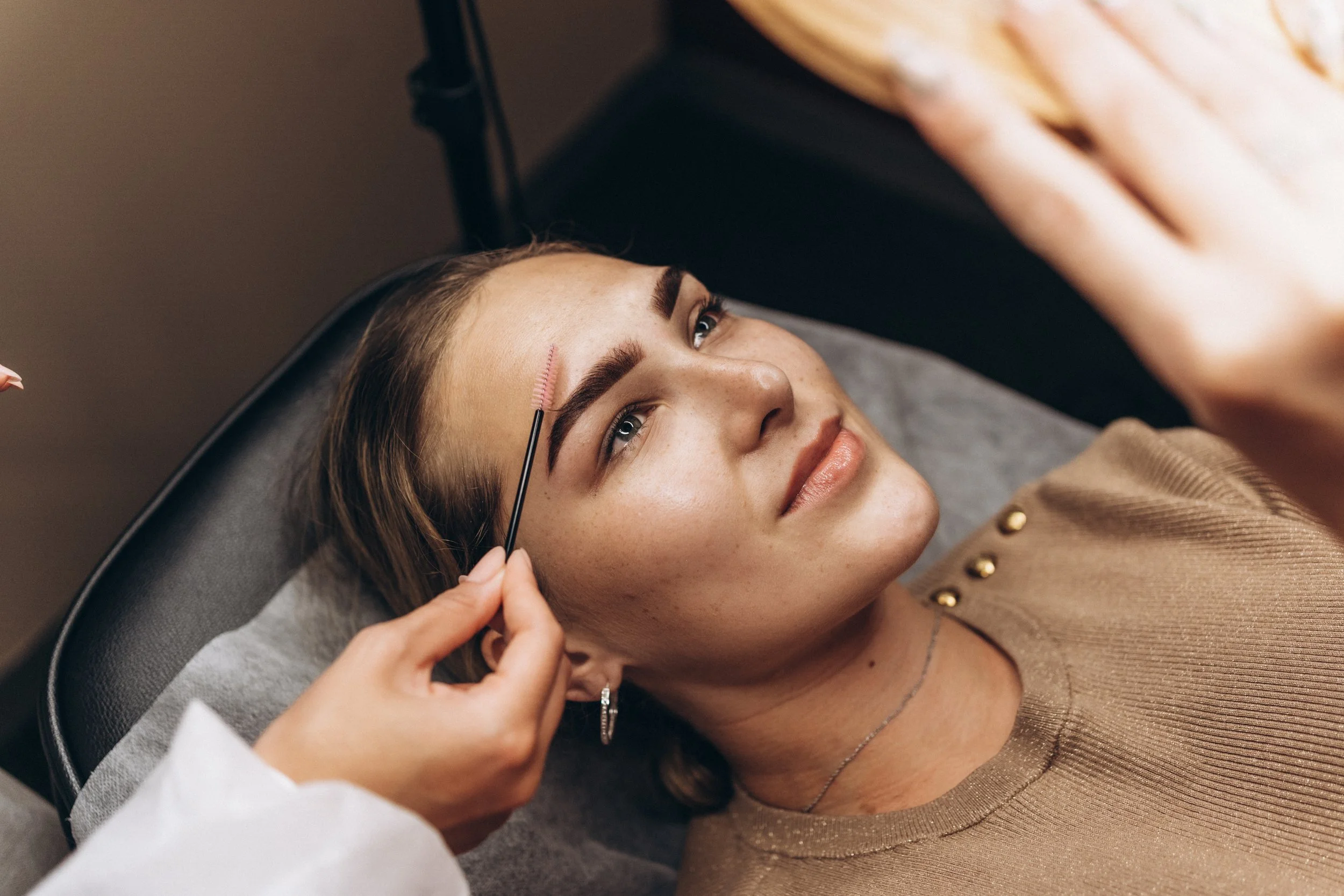 A woman receiving eyebrow grooming at a beauty salon, lying on a chair while a beautician brushes her eyebrows with a small brush.