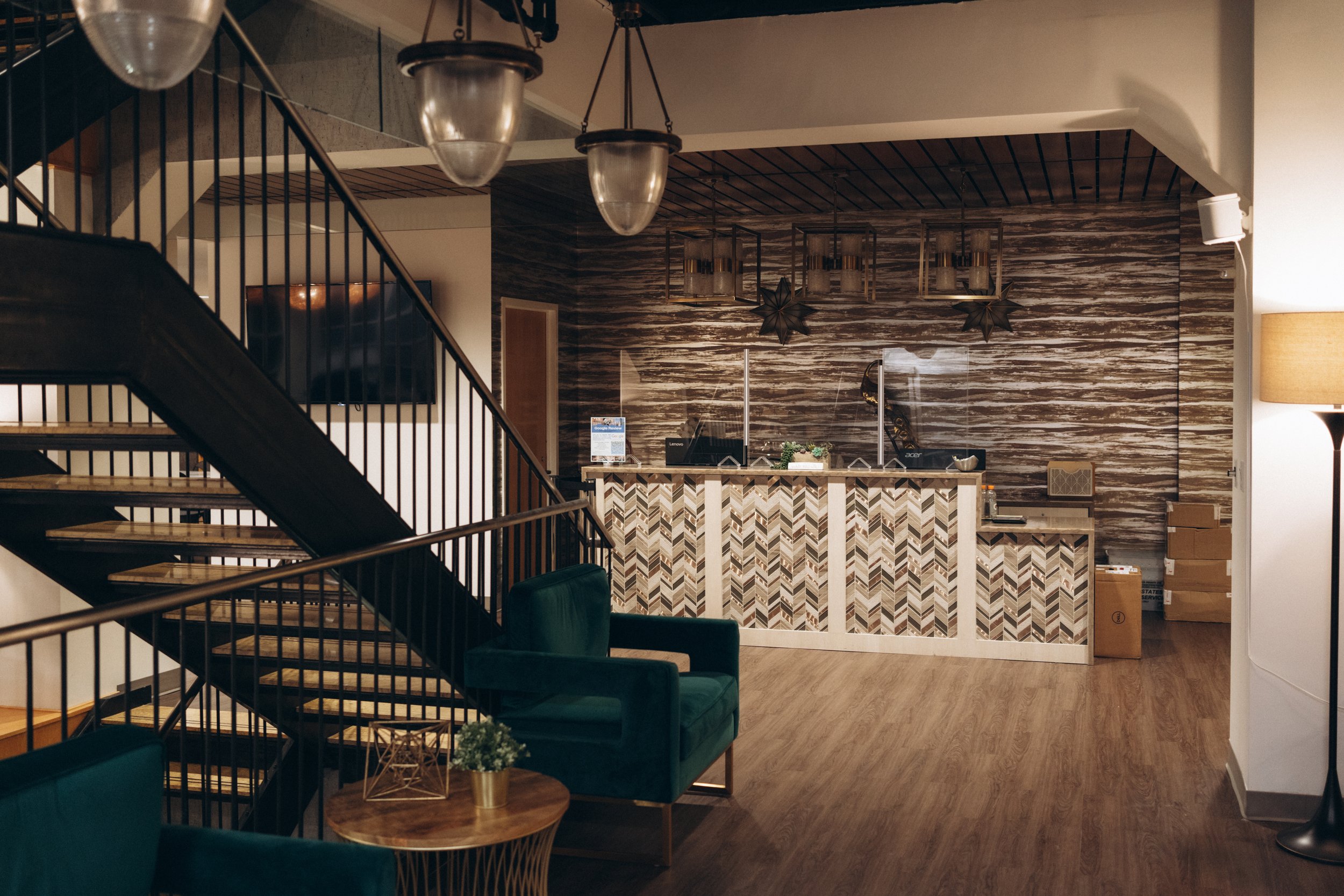 Interior of a hotel lobby or reception area featuring a front desk with a patterned wood design, a staircase with black railings, a large wall with wood paneling, a sitting area with green armchairs beside a small round table with a plant, and a standing lamp.