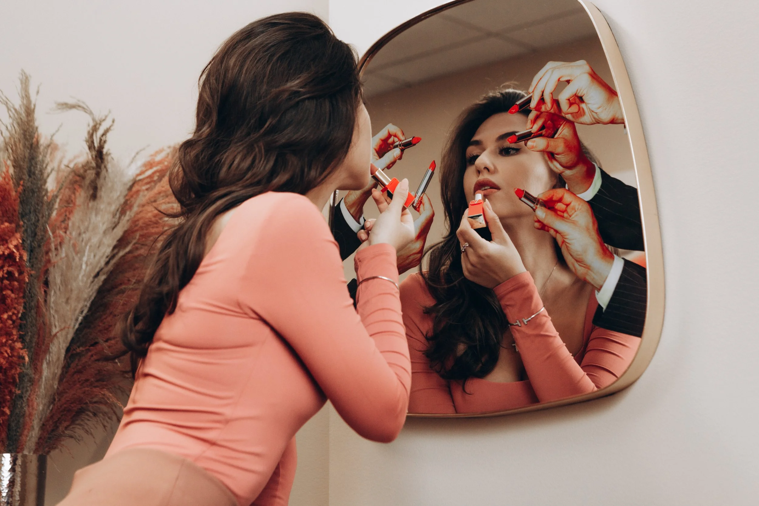 A woman applying lipstick while looking into a mirror, surrounded by multiple hands holding makeup items behind her reflection.