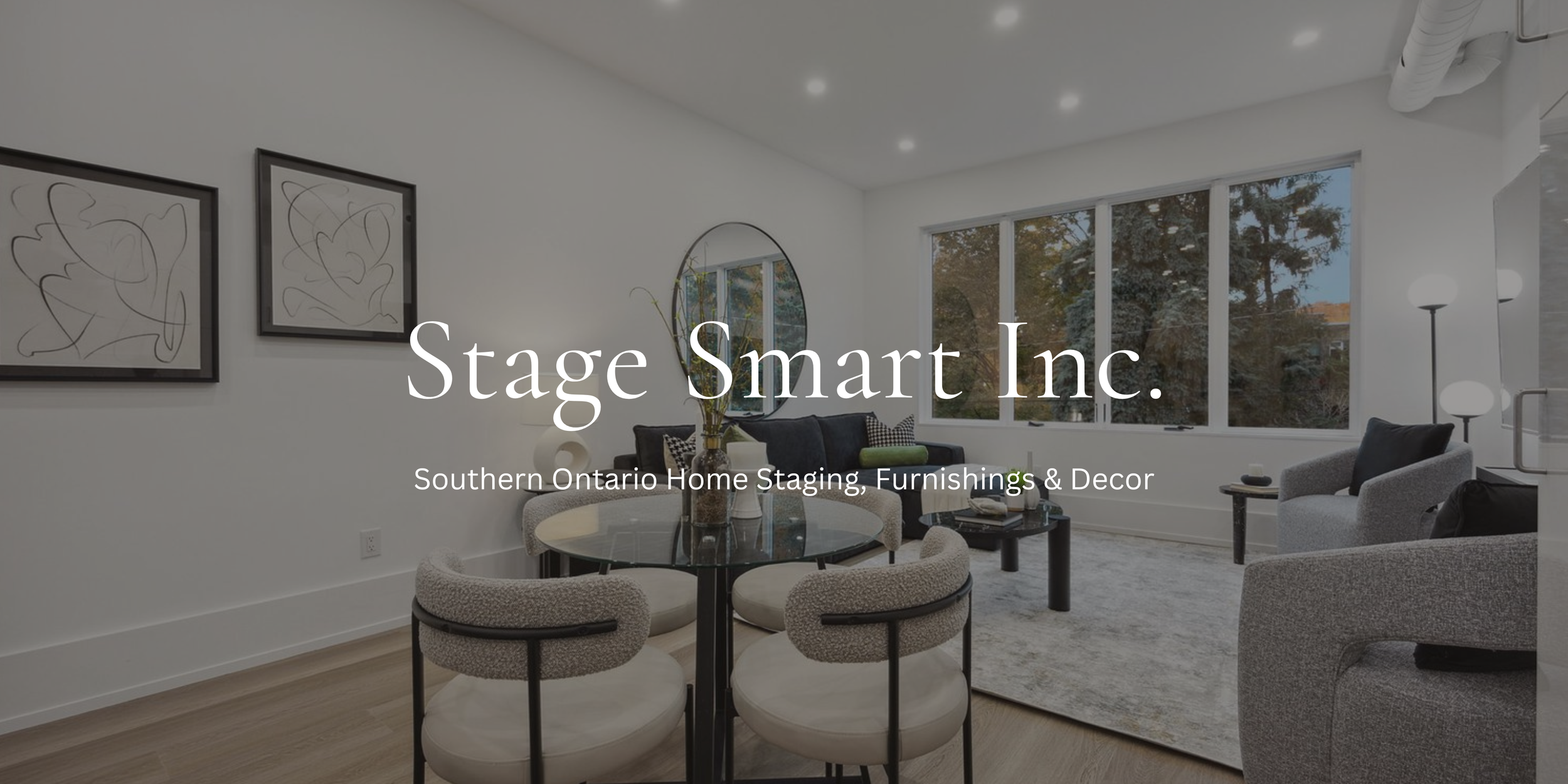 Living room with gray sofas, round glass dining table, large window with trees outside, modern art on white walls, and indoor plants.