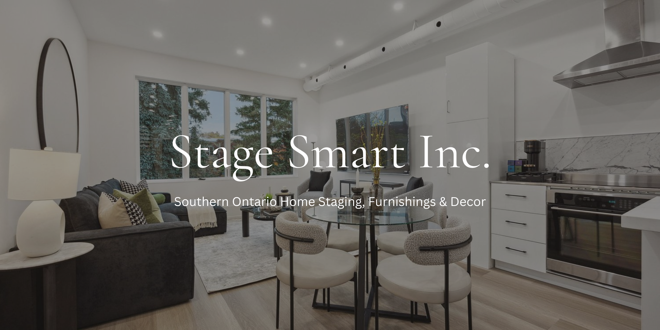 Modern living room with large windows, gray sofa with pillows, round glass dining table with six chairs, TV, and kitchen area with white cabinets and stainless steel appliances.