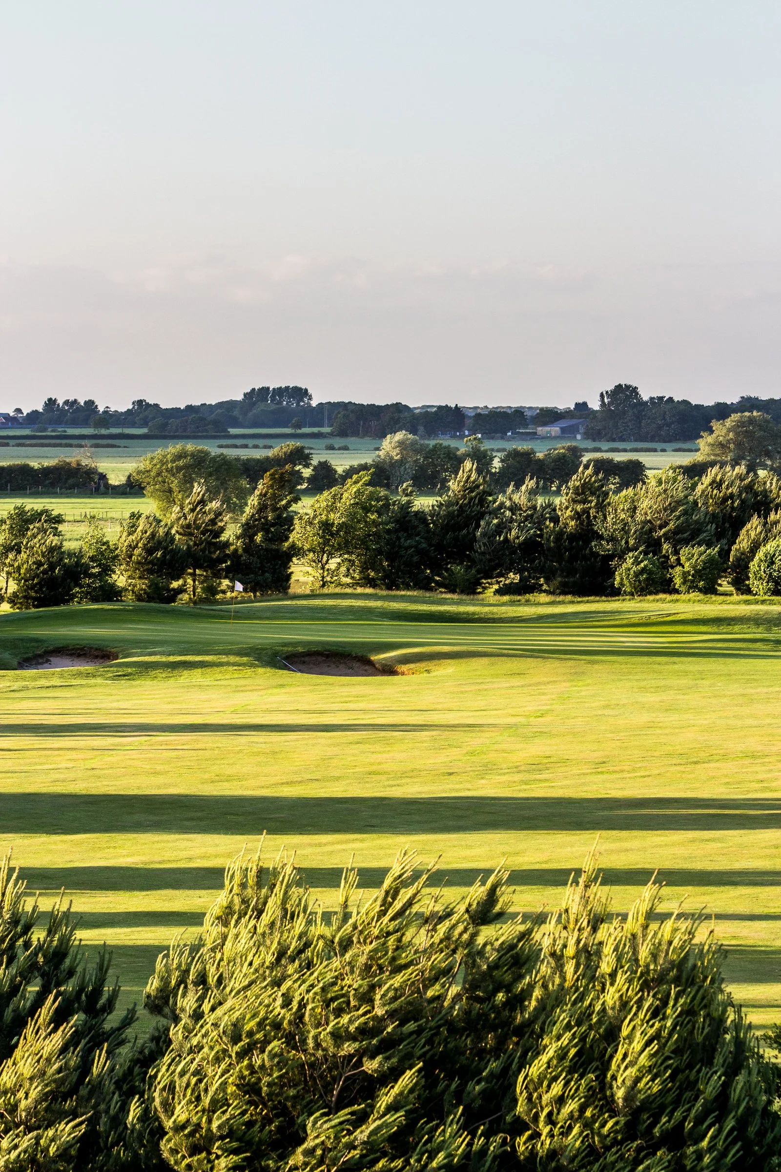 A scenic view of a golf course with green fairways, bunkers, and a flagstick, surrounded by trees and open fields under a clear sky.