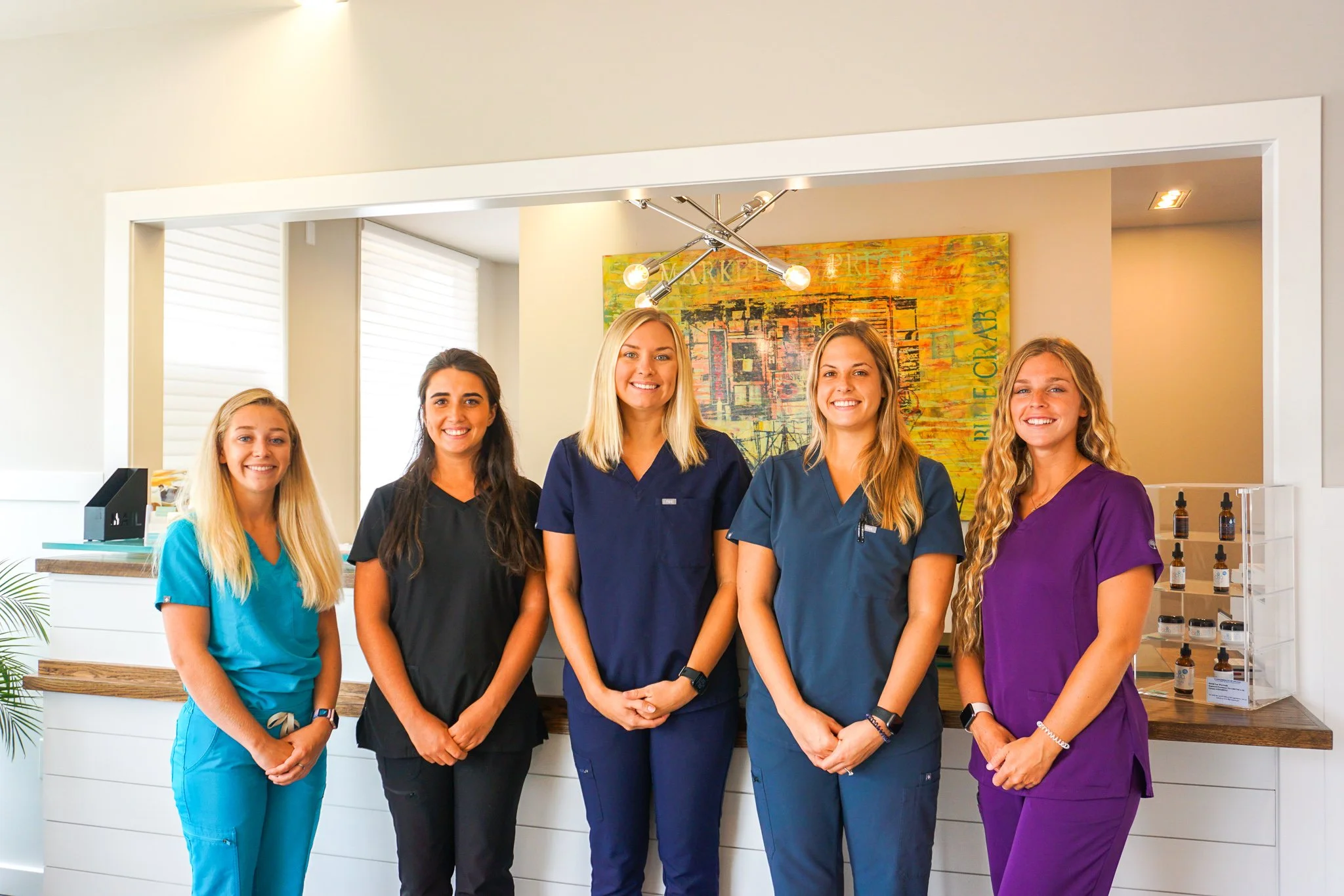 Five women wearing scrubs standing together in a medical or spa reception area smiling at the camera.