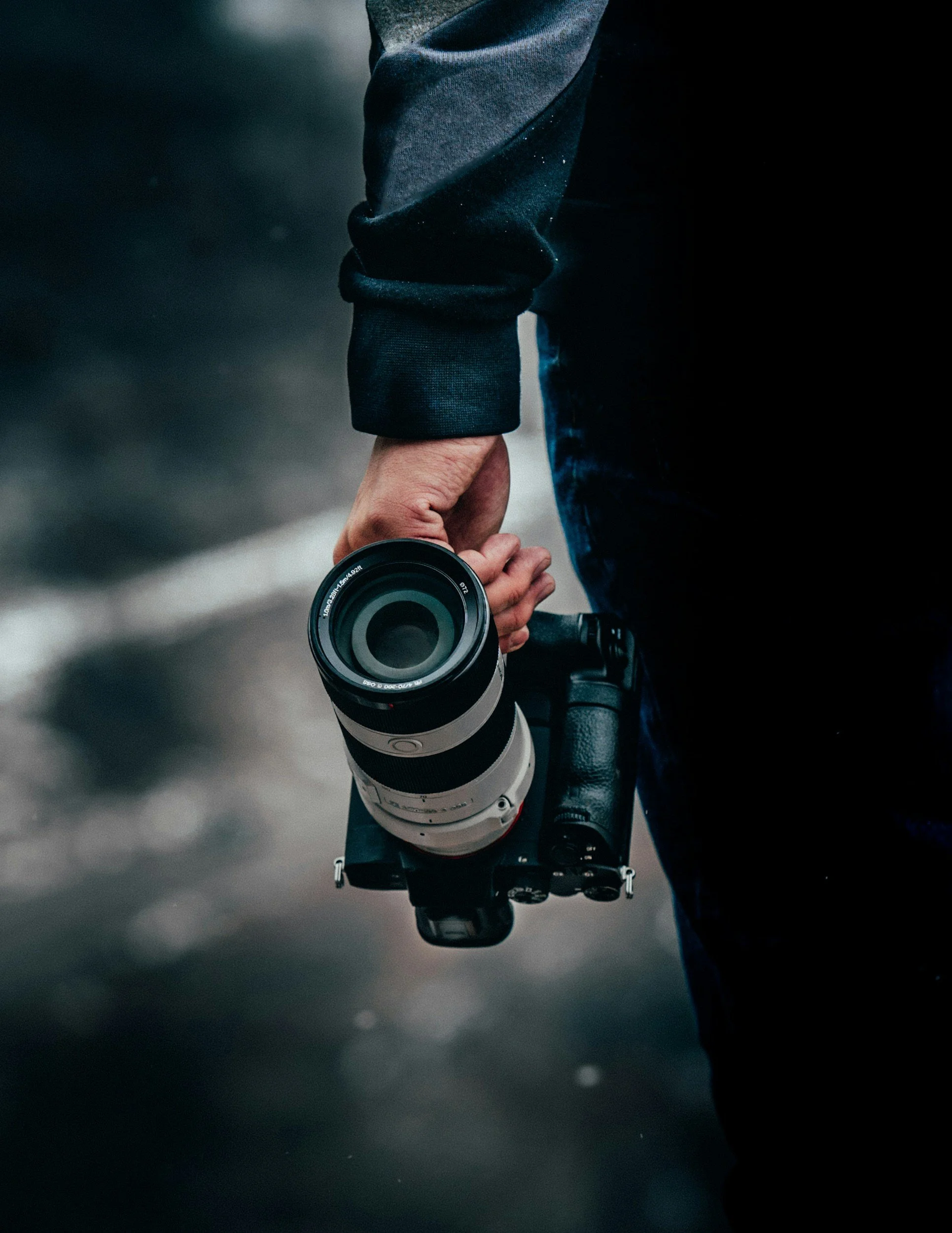 Person holding a camera in their hand, wearing a dark jacket and pants, with the background blurred.