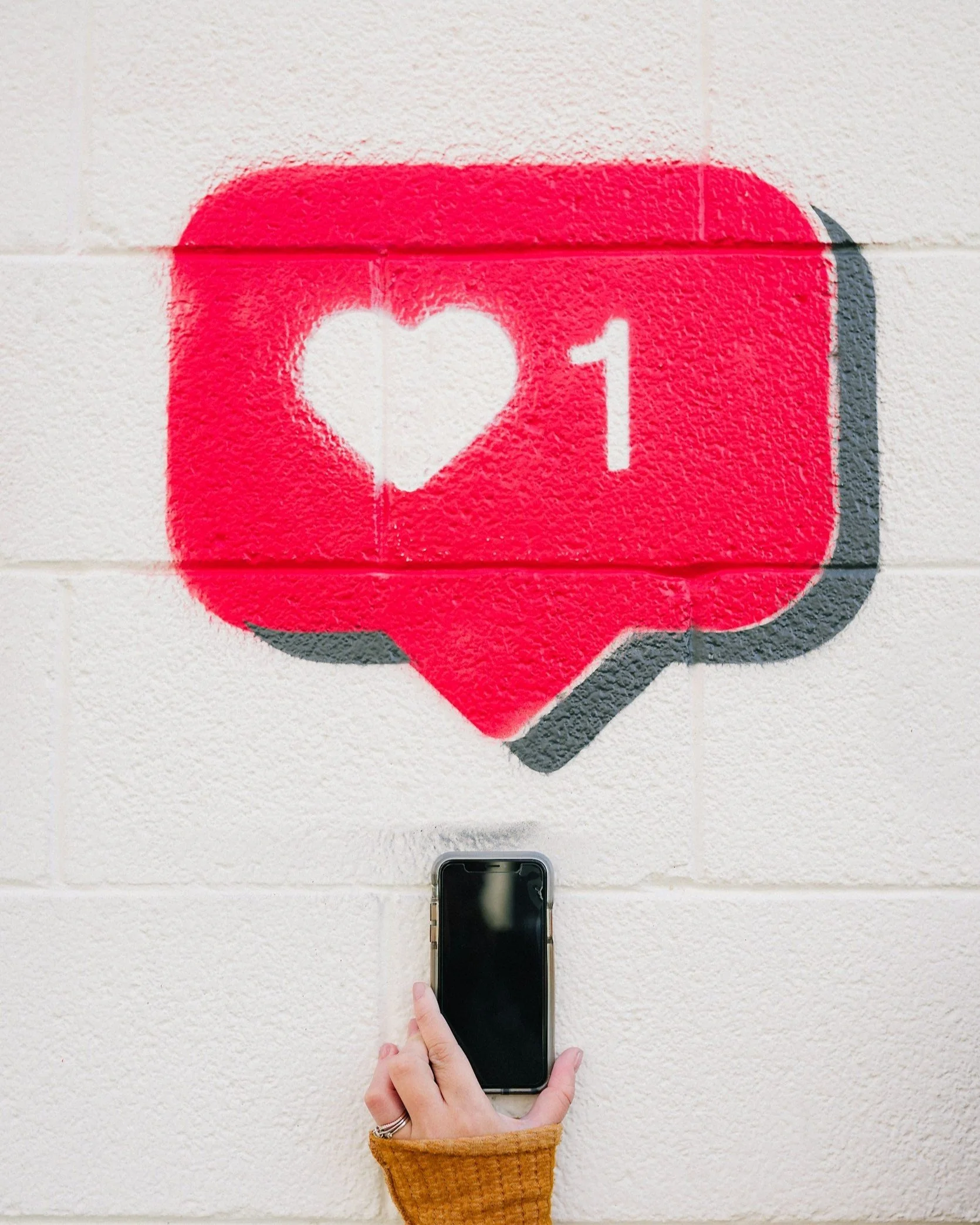 A wall with spray-painted artwork of a red speech bubble with a white heart shape inside and the number 1, resembling a social media like icon. A hand with a wedding ring and a brown sweater points a smartphone at the artwork.