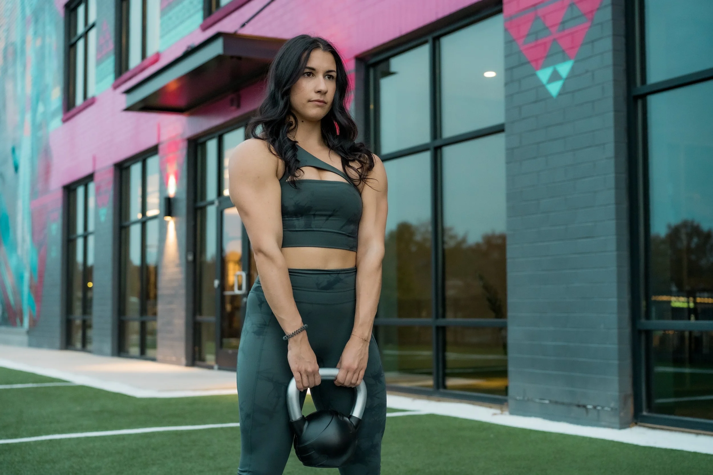 A young woman with long dark hair in athletic wear holding a kettlebell outside a modern building with pink and blue geometric wall art.