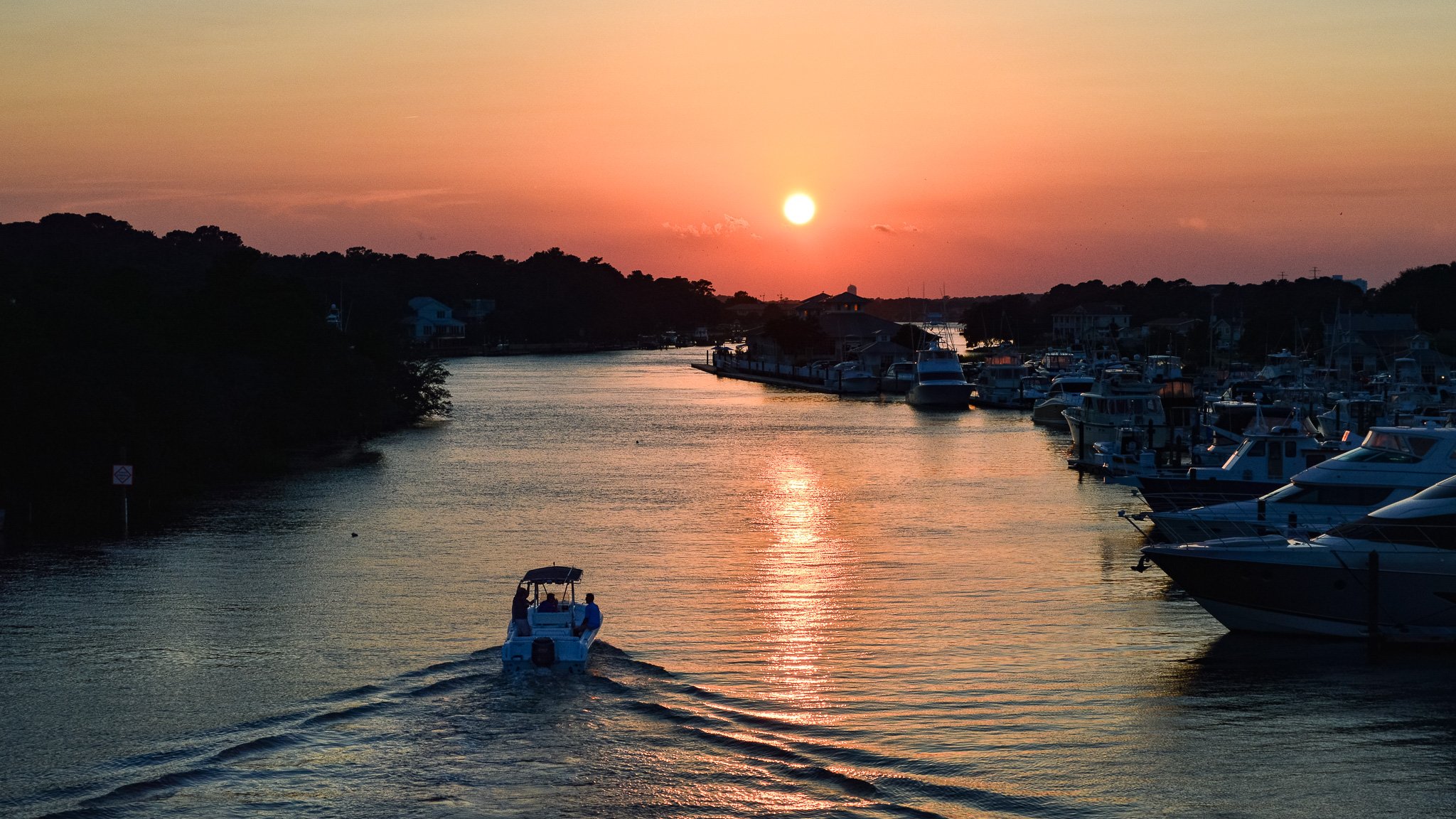 Sunset over a marina with boats docked along the water, a small boat with two people sailing, and a hilly landscape with houses in the background.