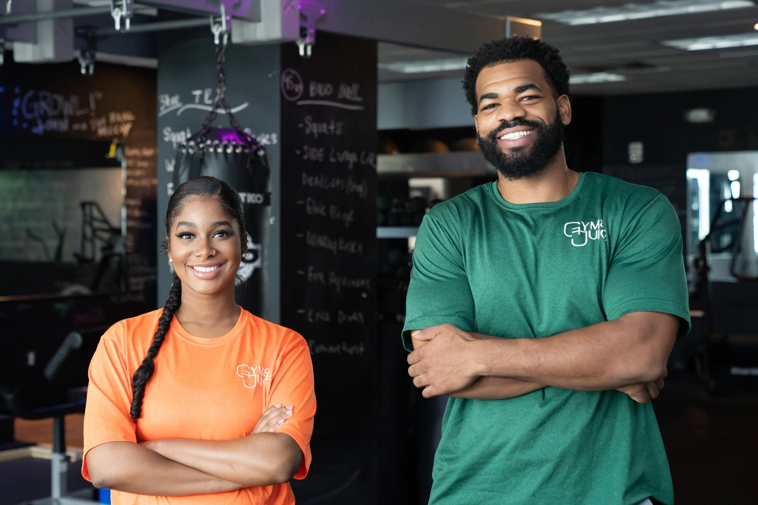 Two smiling individuals standing inside a gym, one wearing an orange shirt and the other wearing a green shirt, with workout equipment and a chalkboard menu in the background.