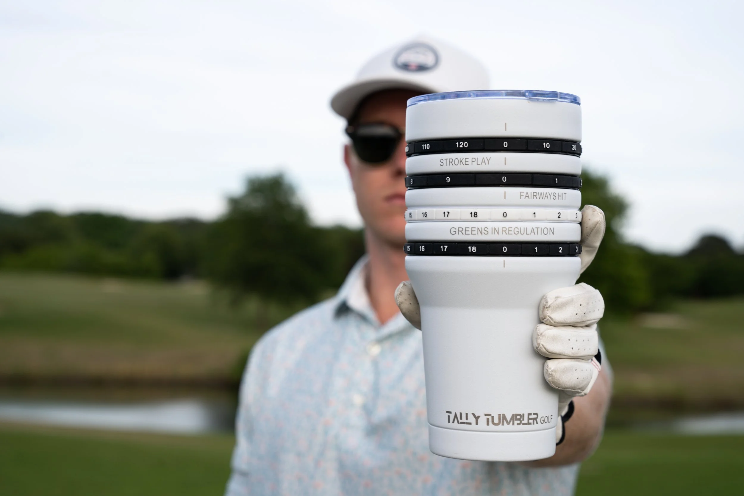Person holding a white Tally Tumbler golf cup with black and white markings, wearing a white glove, a white cap, sunglasses, and a short-sleeve shirt, outdoors with trees and sky in the background.