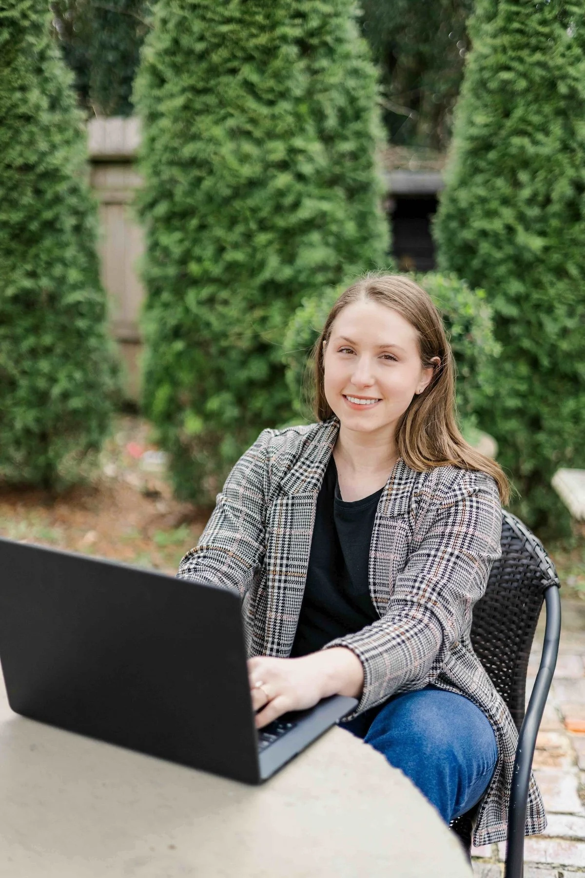 A young woman with light brown hair, wearing a plaid blazer and black top, sitting outdoors at a table with a laptop, smiling at the camera. Green trees and bushes are in the background.