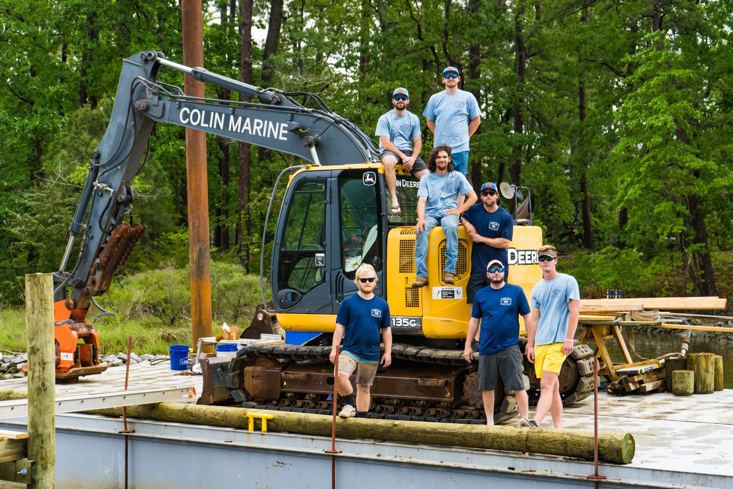 Group of men standing and sitting on a yellow John Deere excavator on a construction site near a body of water, with lush green trees in the background.