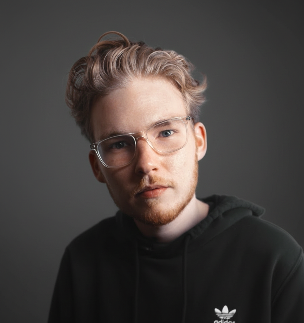 A young man with wavy, blond hair, wearing large clear glasses and a black hoodie with a small white logo, looking directly at the camera against a plain gray background.