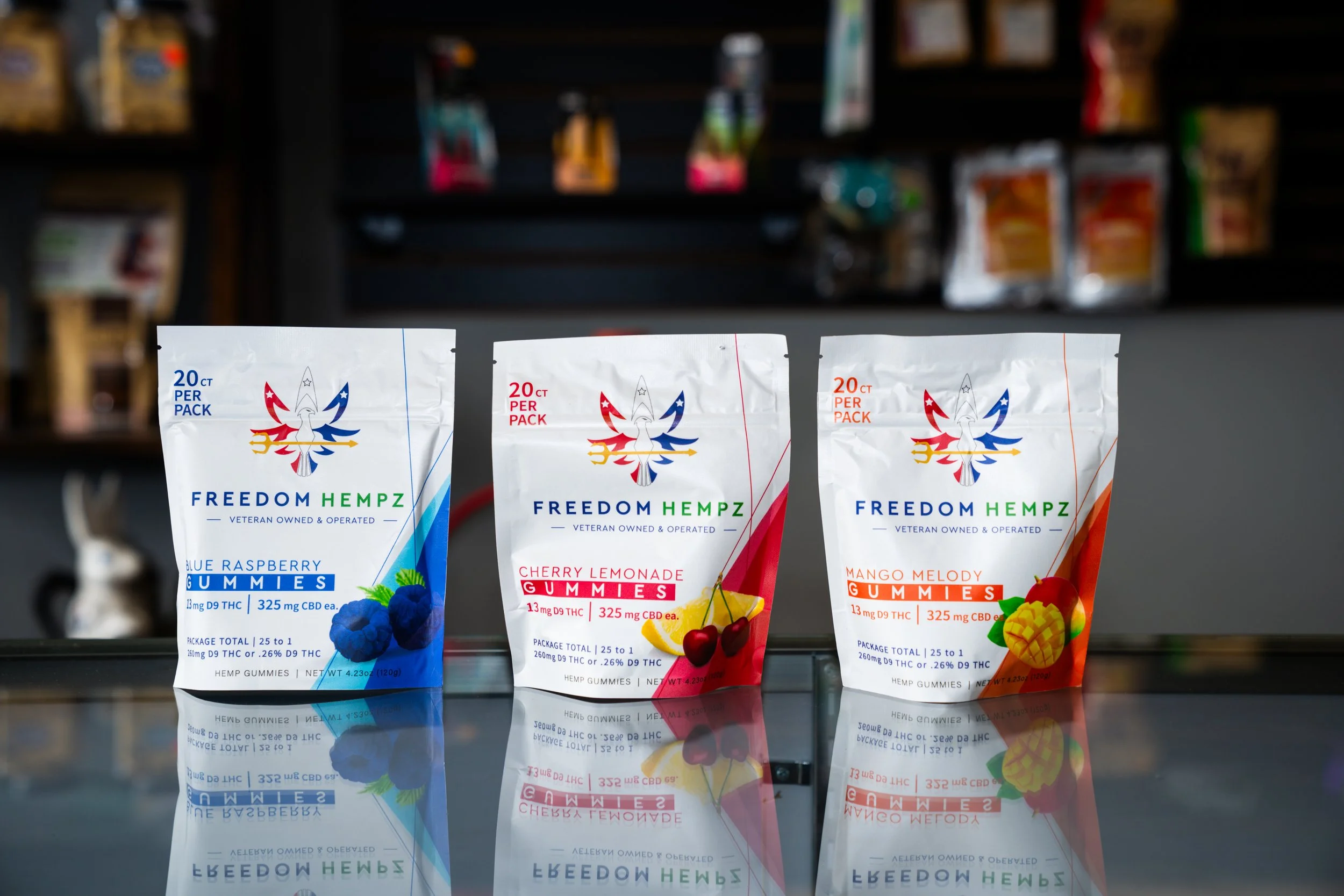 Three packages of hemp gummies displayed on a reflective surface with a dark background. The packages are labeled as blueberry raspberry, cherry lemonade, and mango melody flavors.