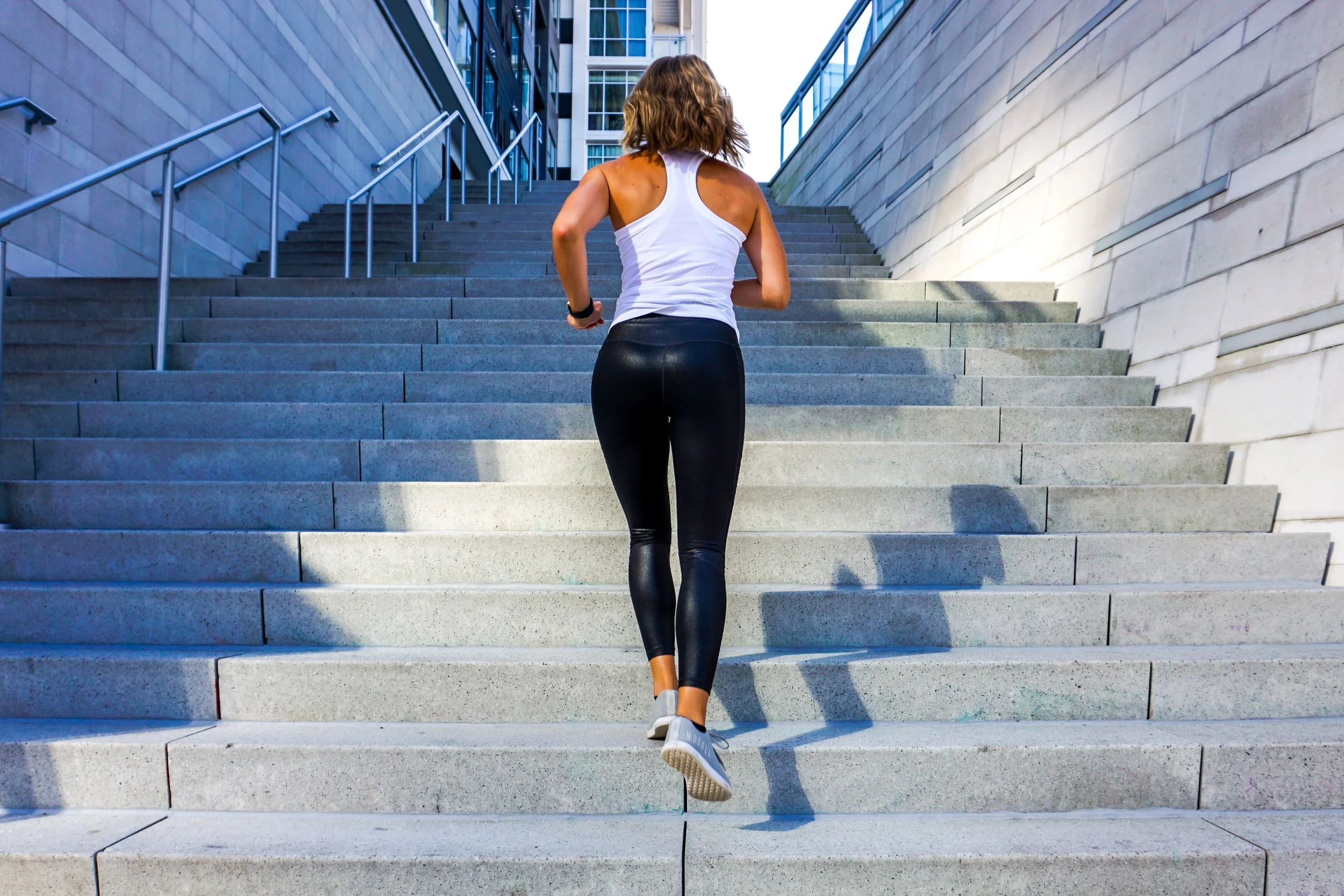 A woman in athletic workout clothing running up an outdoor staircase between modern buildings.