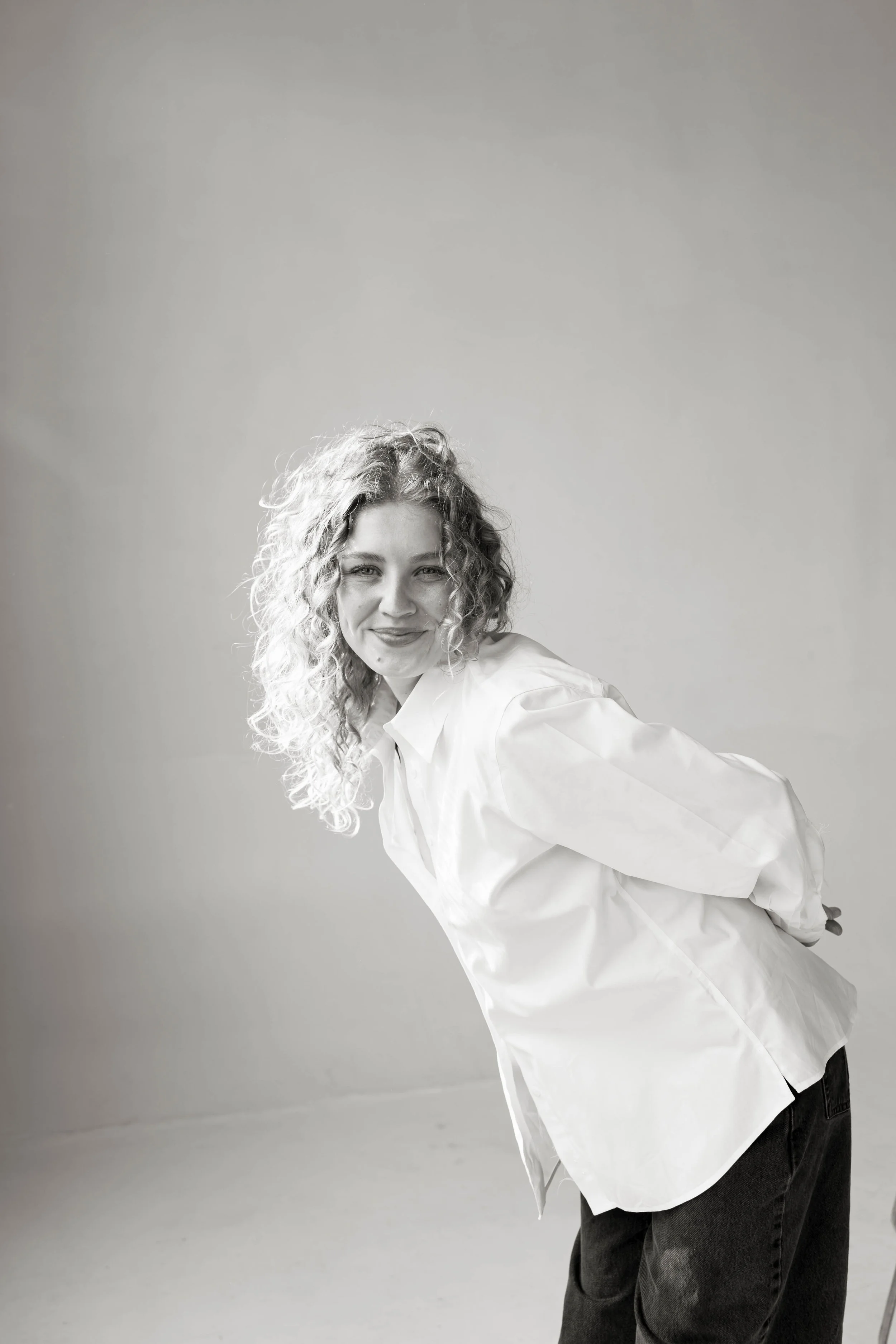A person with curly hair partially bent over, wearing a white shirt and dark jeans, smiling in front of a plain background.